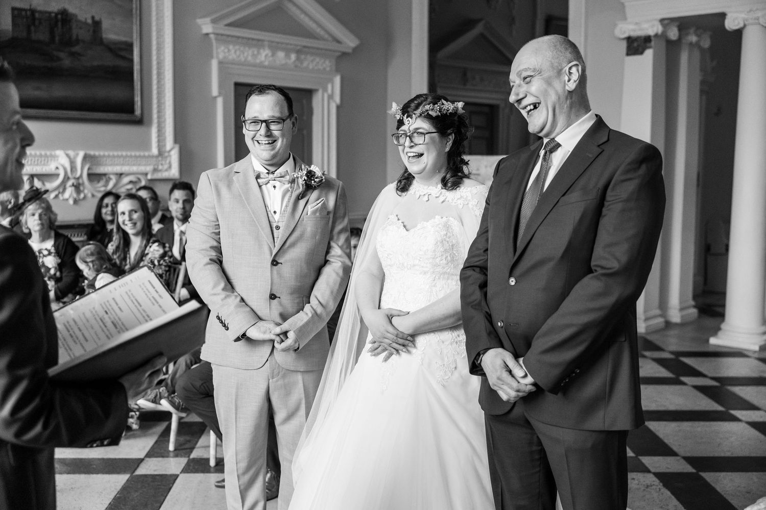 Black and white wedding ceremony photo of smiling bride and groom standing with officiant before seated guests.