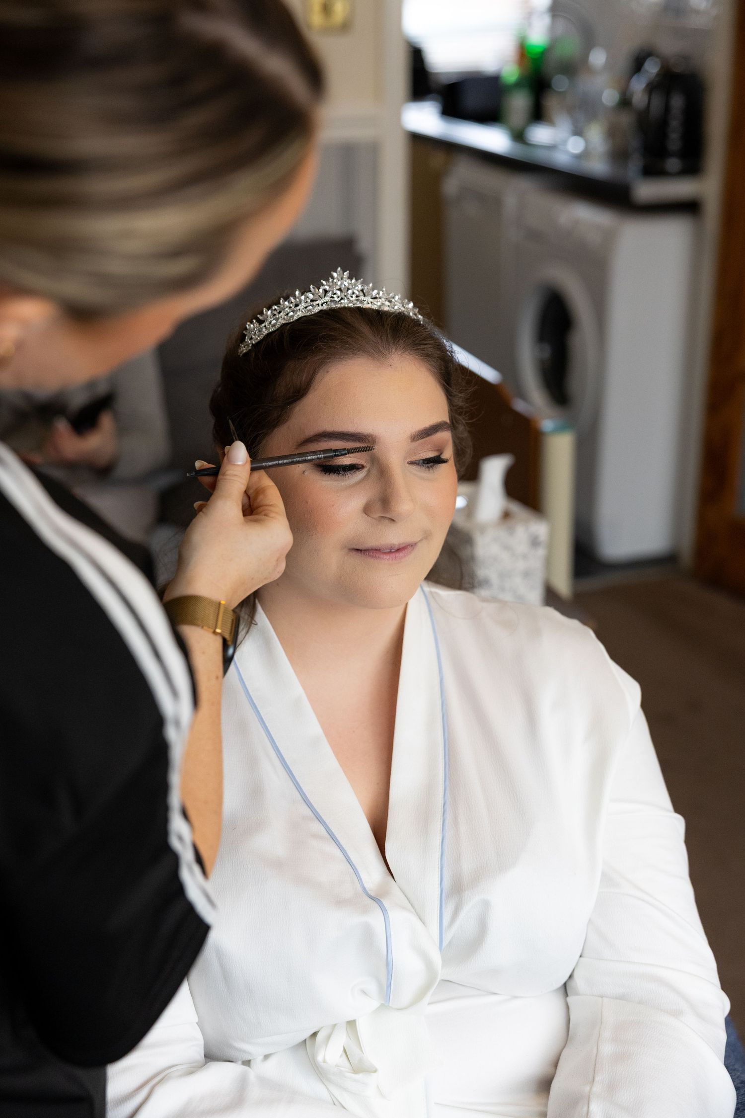 A makeup artist applies eyeliner to a bride wearing a tiara and white robe while getting ready for her wedding.