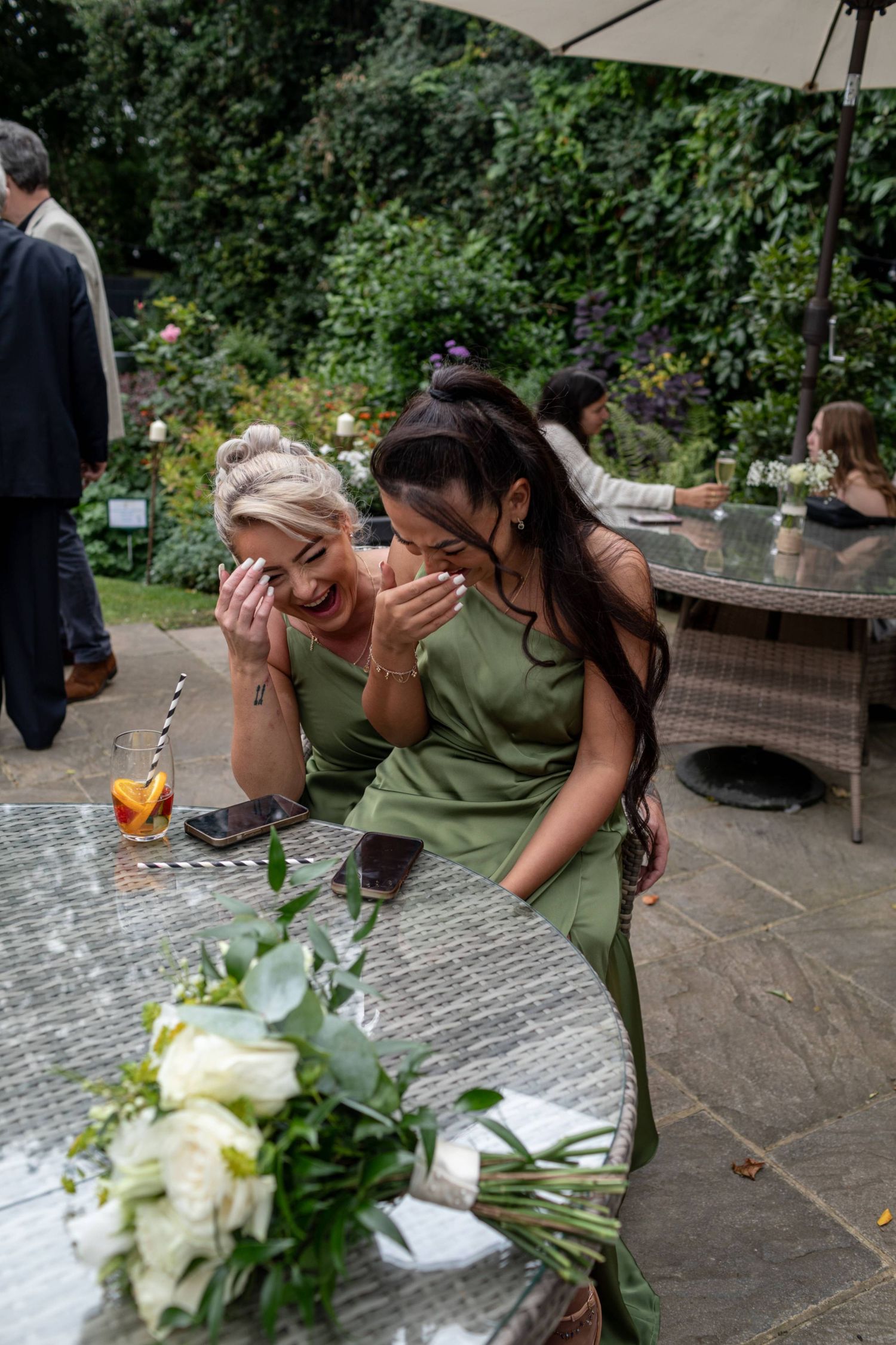 Two bridesmaids in green dresses laughing together at an outdoor wedding reception table with white flowers.