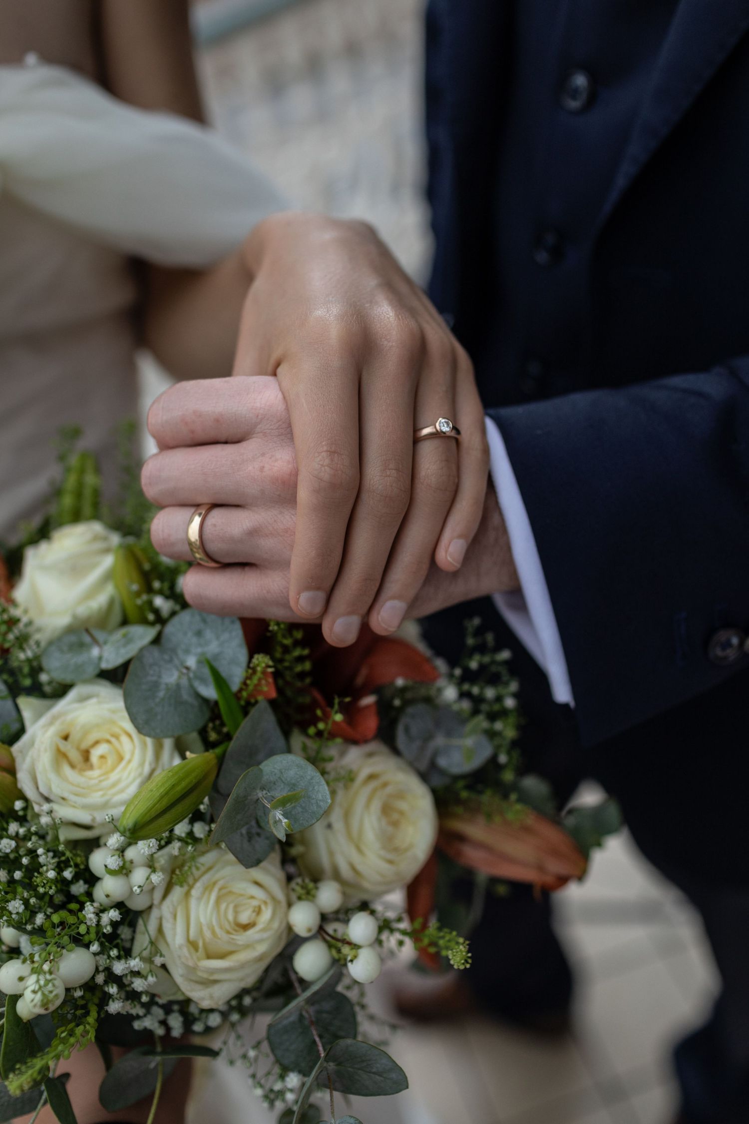 Newlywed couple displaying wedding rings on bouquet of white roses and eucalyptus, groom in navy suit.
