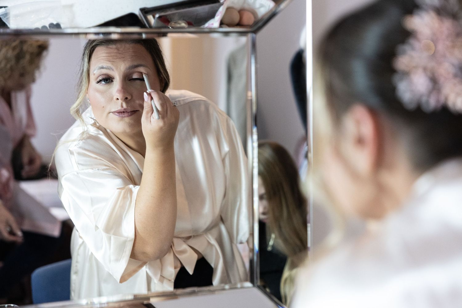 Woman applying eye makeup while looking in a mirror, with others visible in the background.