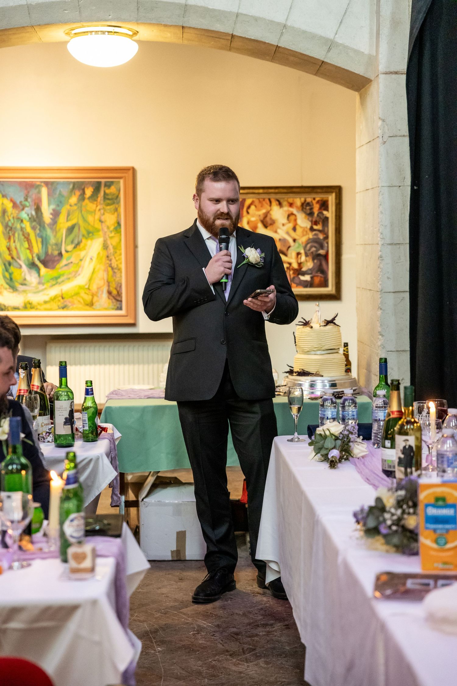 Groom in black suit giving wedding speech at reception, holding microphone and phone, with decorated tables and artwork behind.