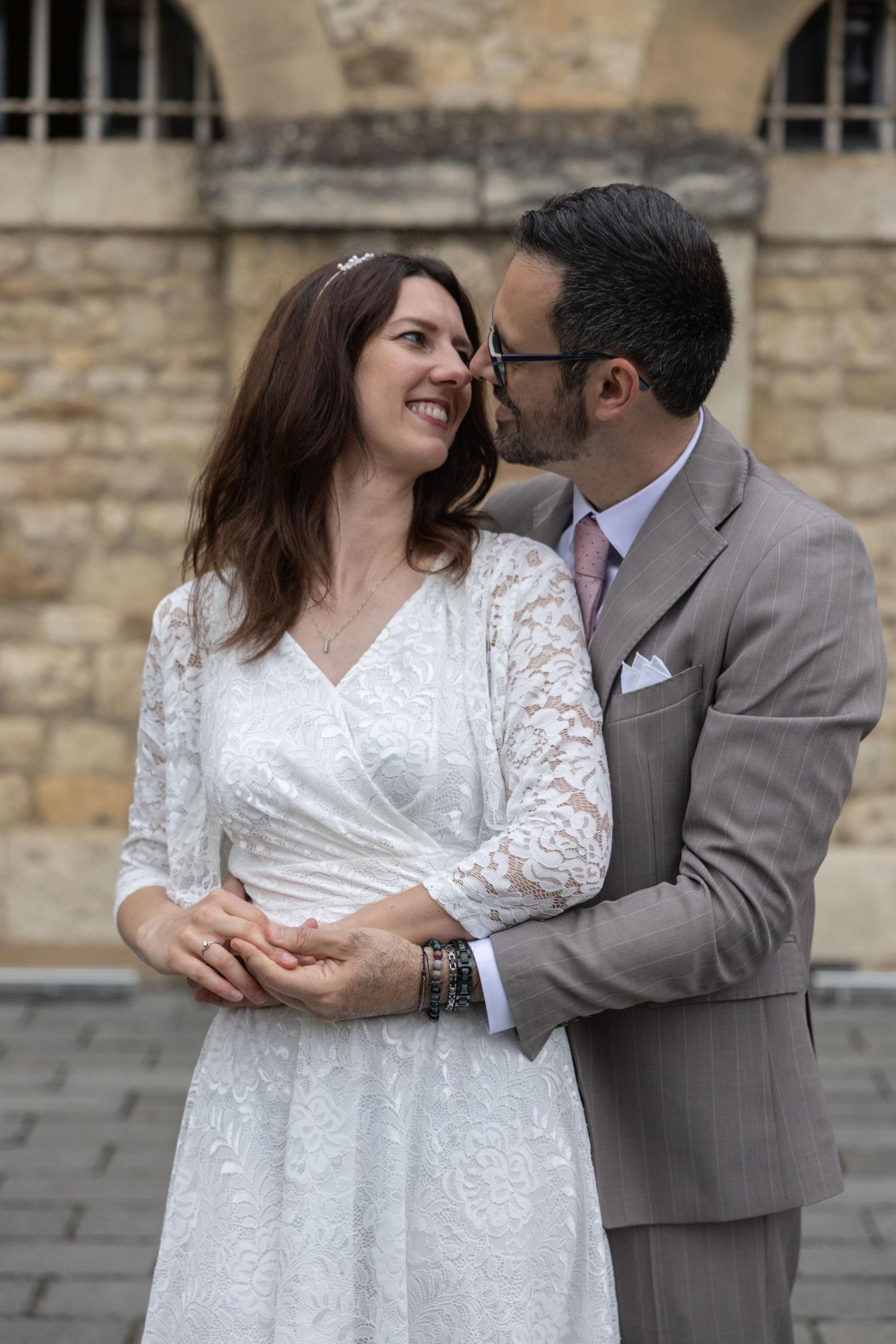 A bride in white lace dress and groom in gray suit share a romantic moment, smiling at each other outdoors.