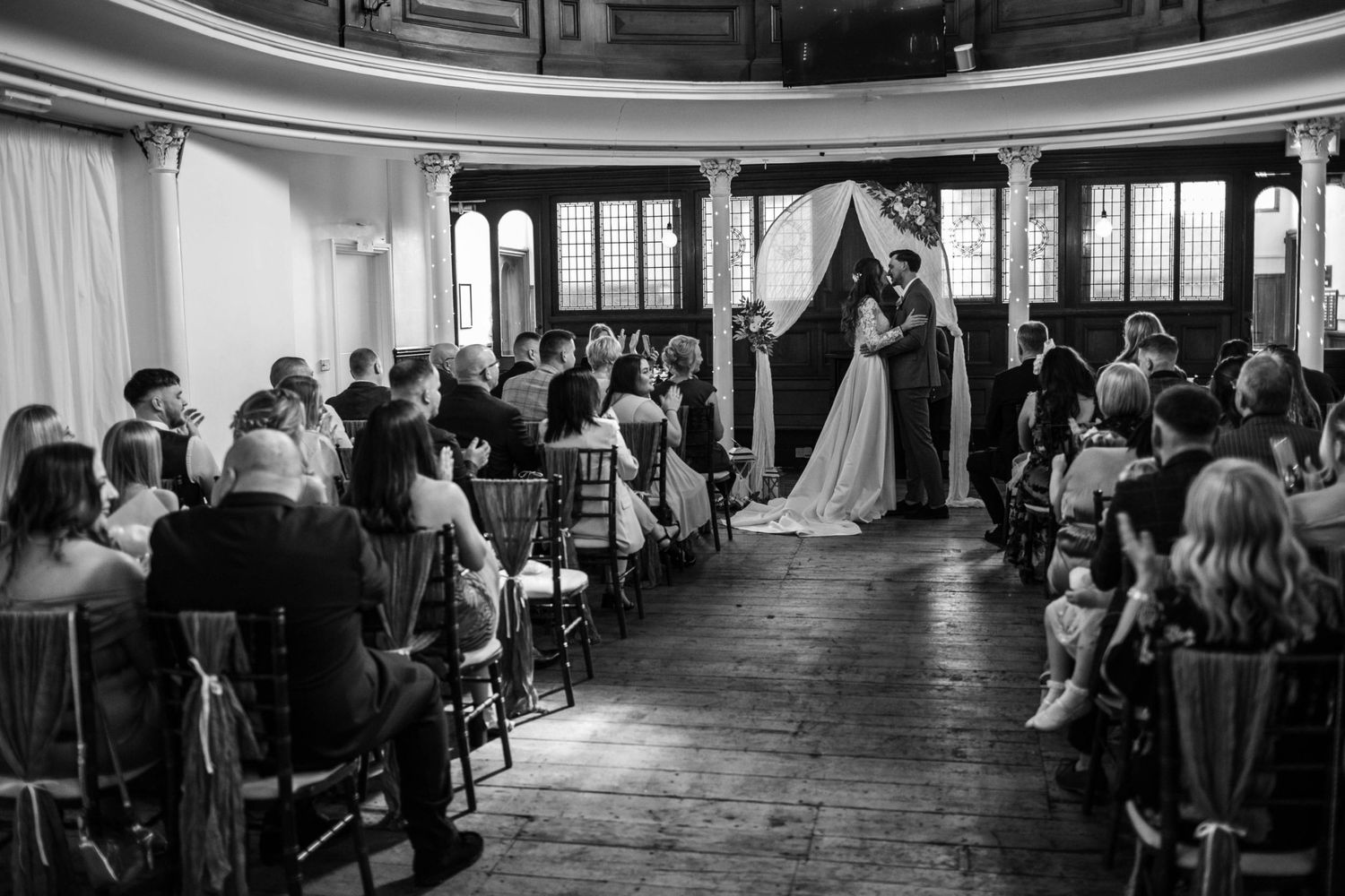 Black and white wedding ceremony photo with couple at altar, guests seated in elegant venue with wooden floors.