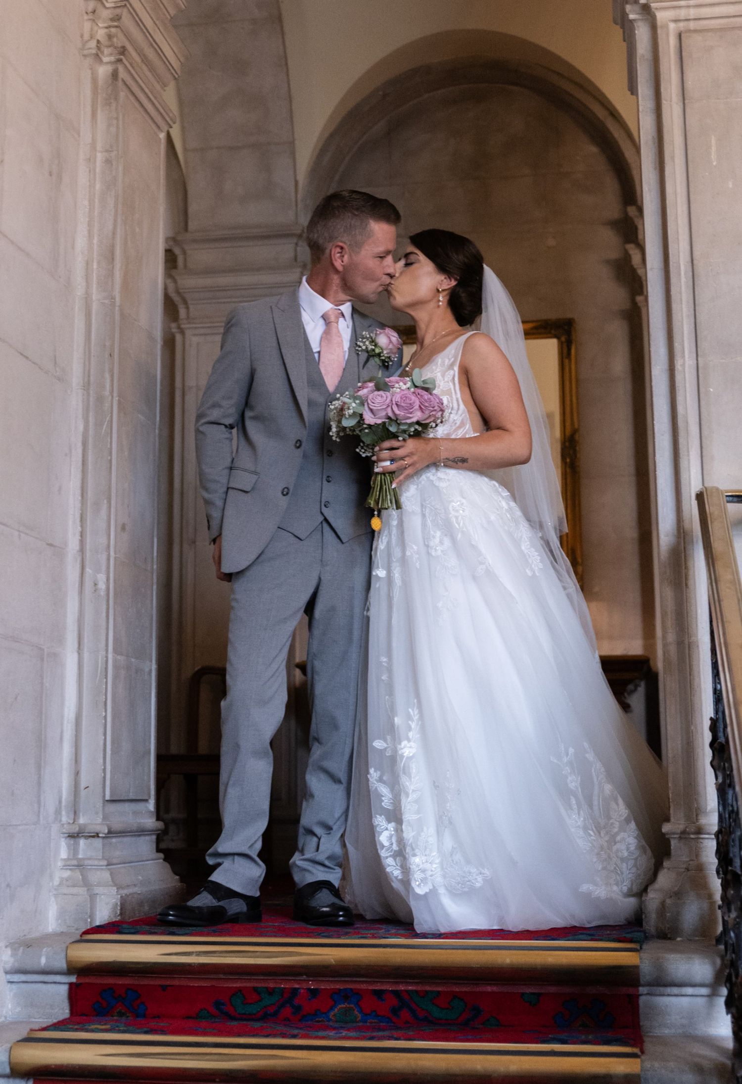 Bride and groom share a romantic kiss on grand staircase, bride holding pink roses bouquet in elegant white gown.