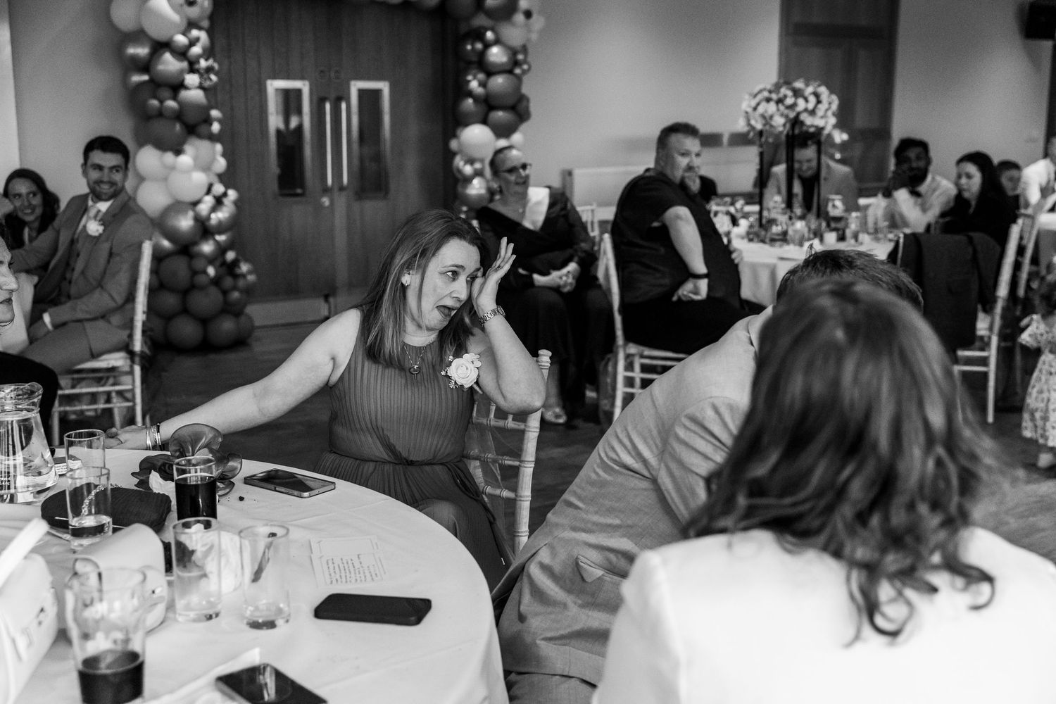 Black and white photo of guests socializing at an elegant event with balloon arch decor and round tables.