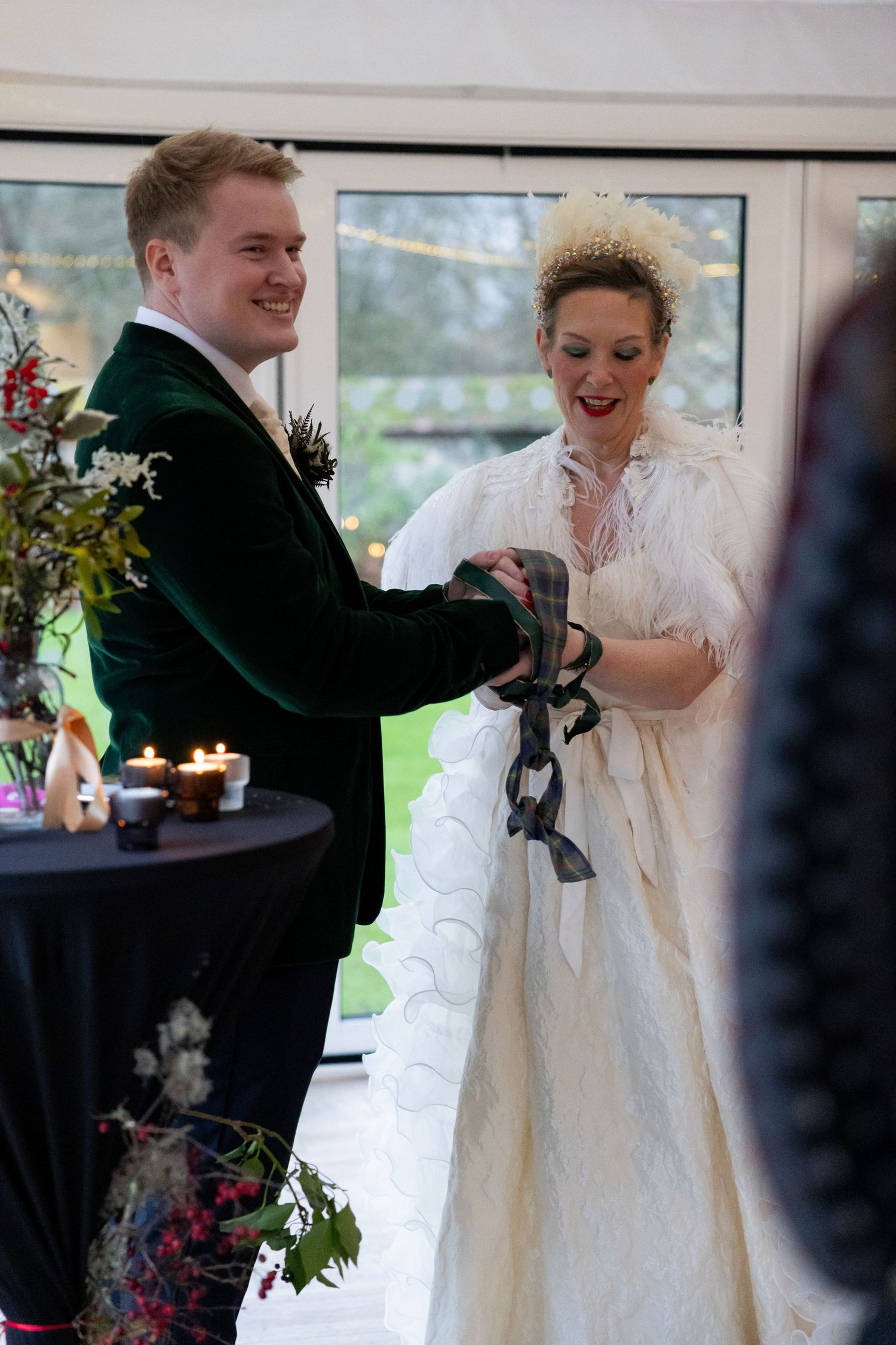 A couple in formal wedding attire share a hand-bonding moment in a bright indoor venue.