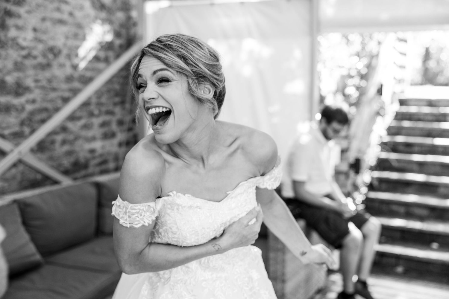 Joyful bride in off-shoulder lace wedding dress laughing exuberantly in black and white photo.