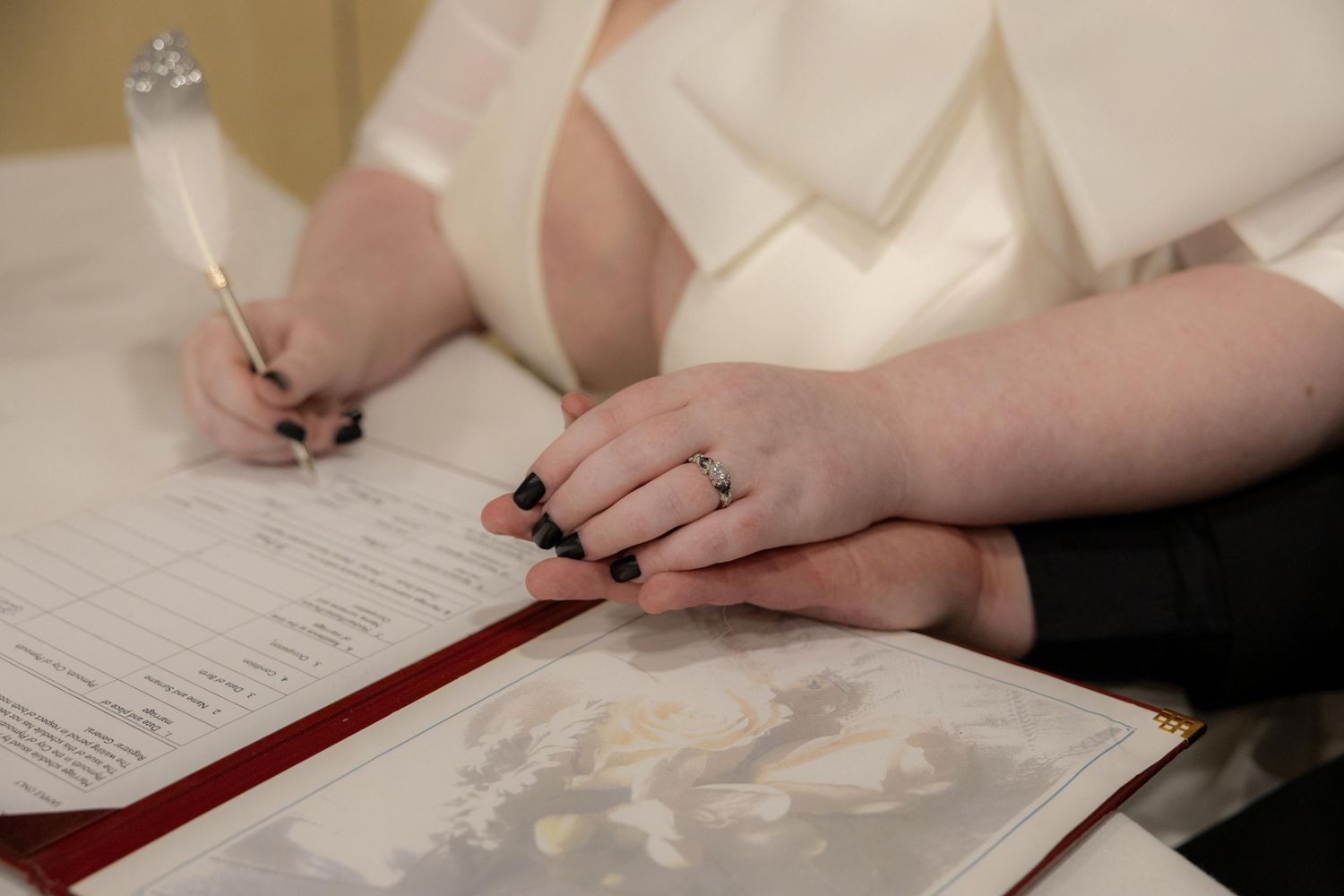 Bride signing wedding registry documents with black nail polish, wearing engagement ring, holding hands with groom.