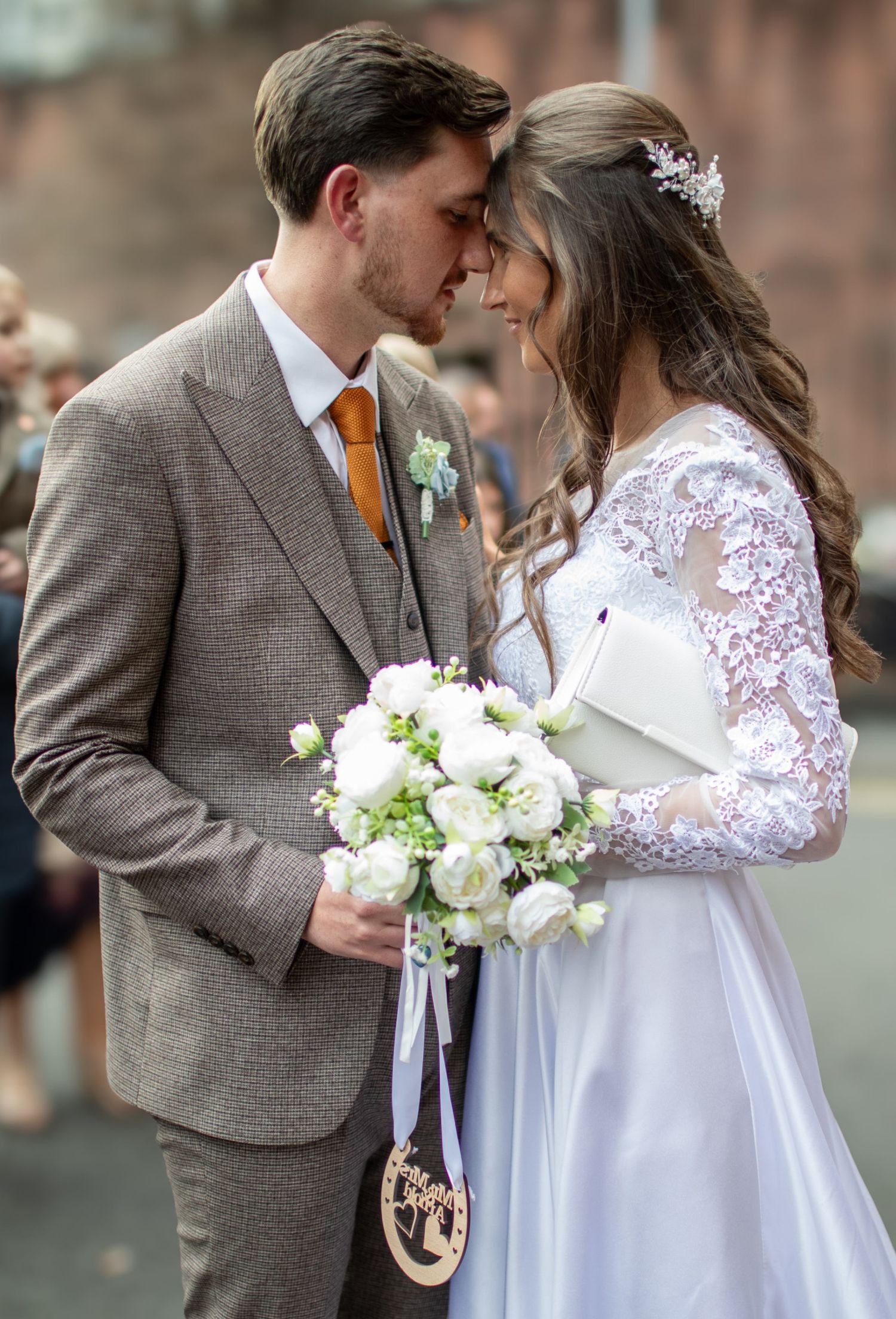Bride and groom share a romantic moment, she holds white rose bouquet, wearing lace dress and floral crown.