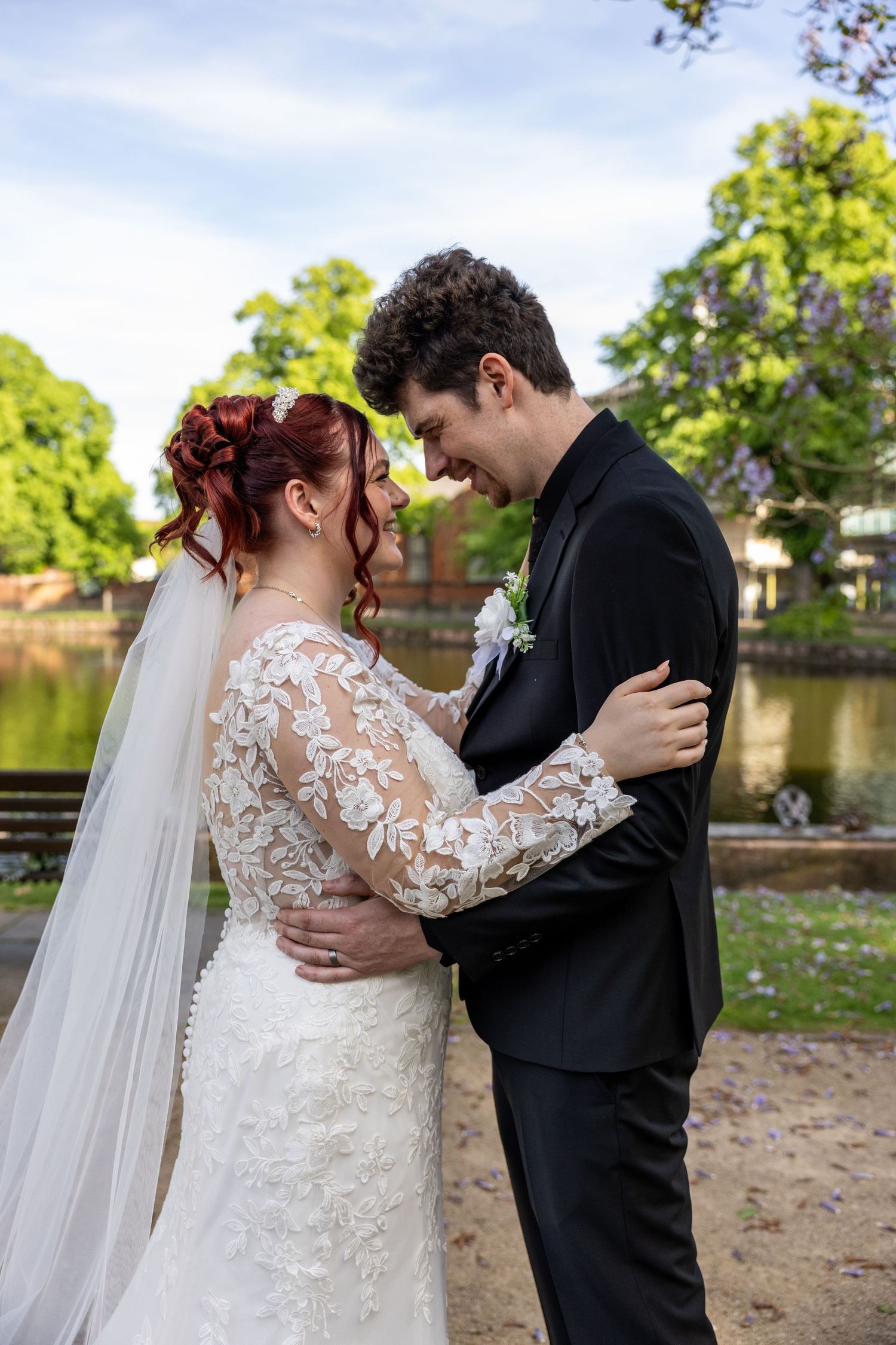 Bride in lace gown and groom in black suit embrace and smile at each other in a park setting near a pond.