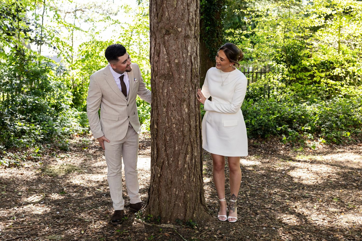 A bride in a white dress and groom in a beige suit stand on opposite sides of a tree in a sunlit forest.
