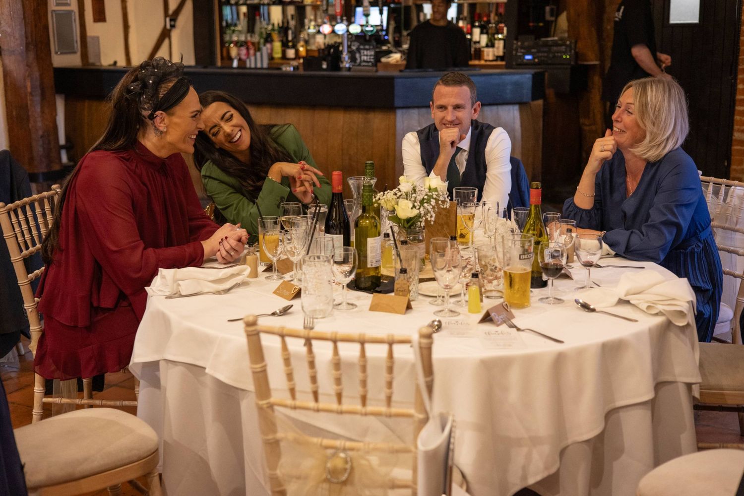 Four people enjoy a lively dinner at a restaurant table with wine bottles and floral centerpiece.