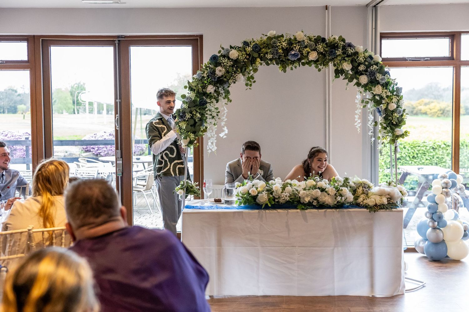 Wedding reception speech with floral arch backdrop, couple seated at head table with white flower arrangements.
