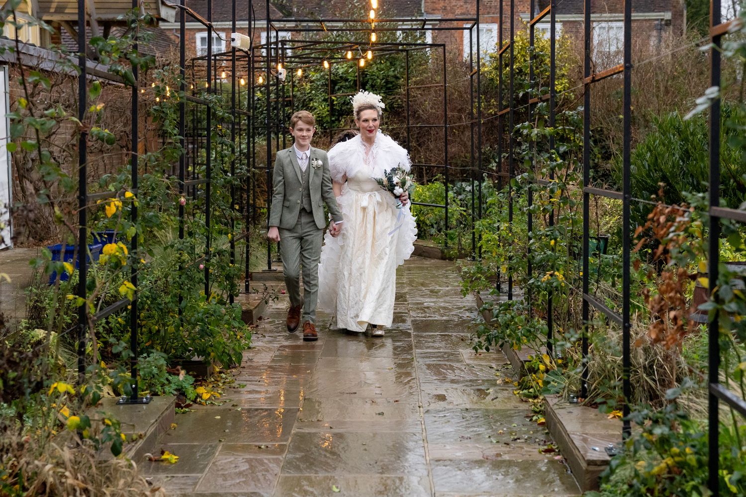 People walk through a lush garden pathway with arched trellises covered in climbing plants and flowers.