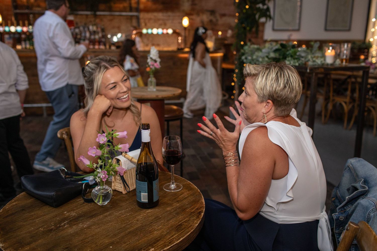 Two women laughing and conversing over wine at a rustic wedding reception venue.