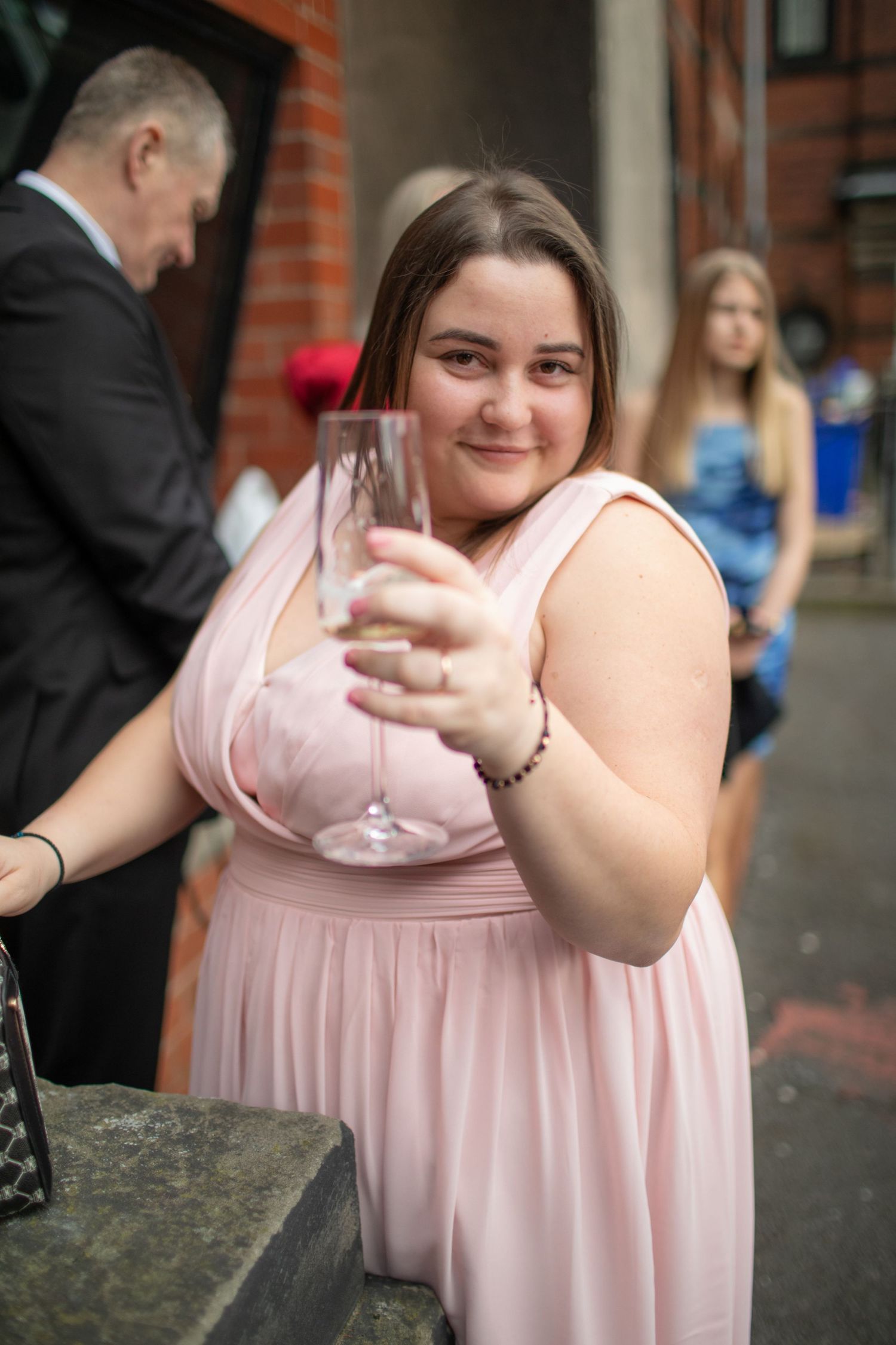 Young woman in pink dress holding champagne glass at formal event, smiling at camera outdoors.