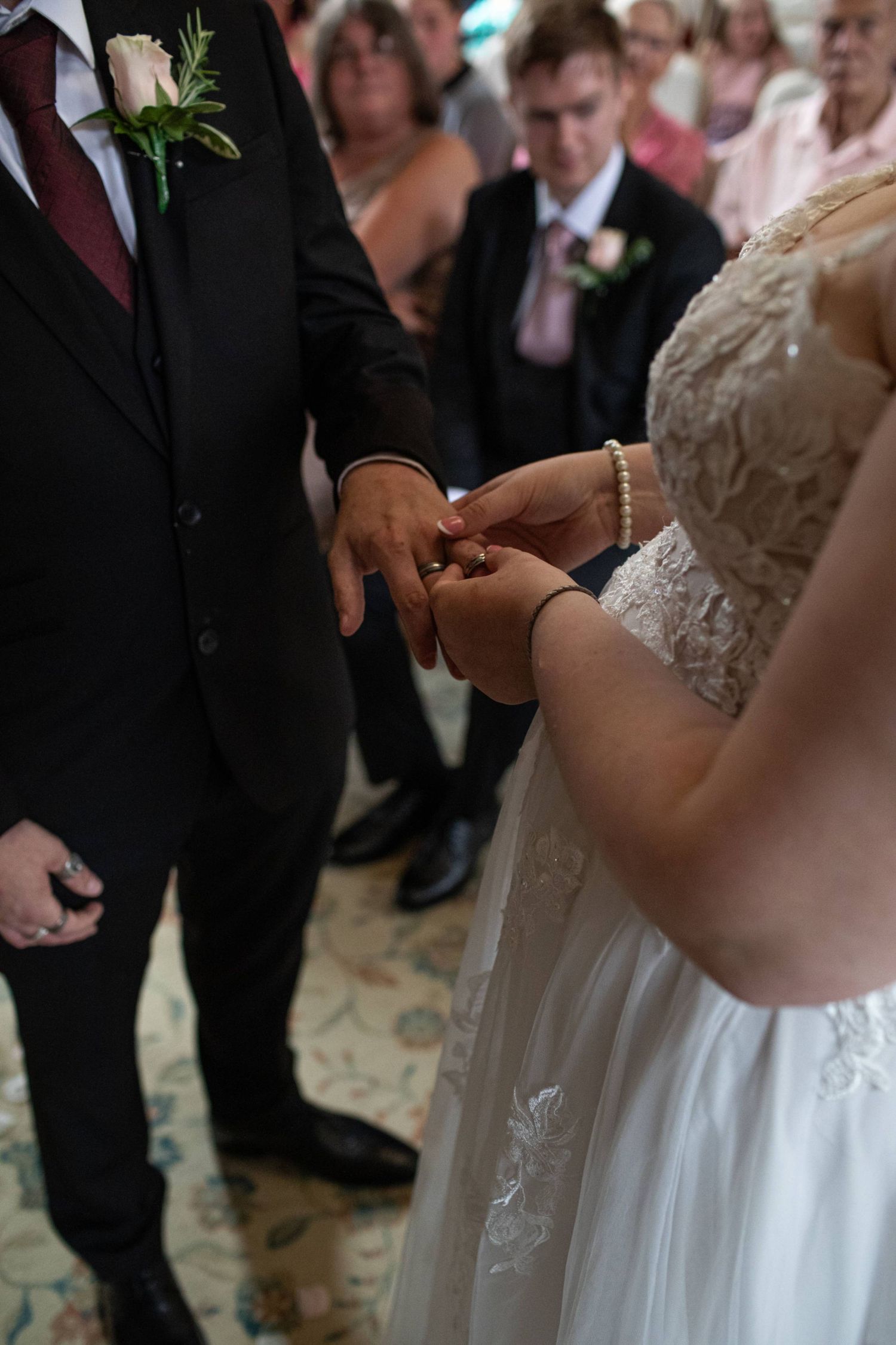 Bride placing wedding ring on groom's finger during romantic ceremony with guests watching in background.
