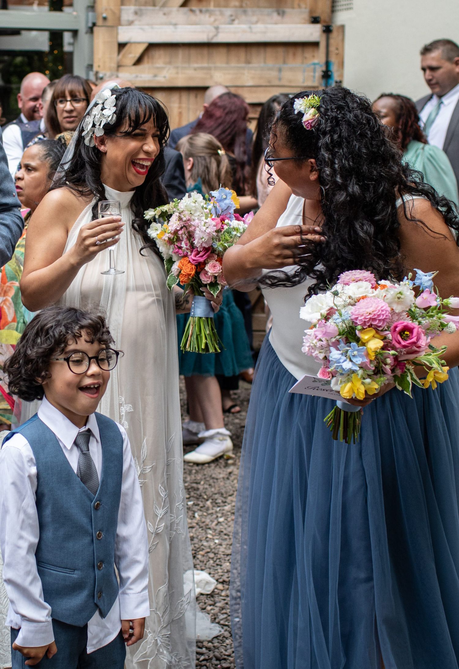 Bride in white gown laughs with bridesmaid holding colorful bouquets at outdoor wedding ceremony.