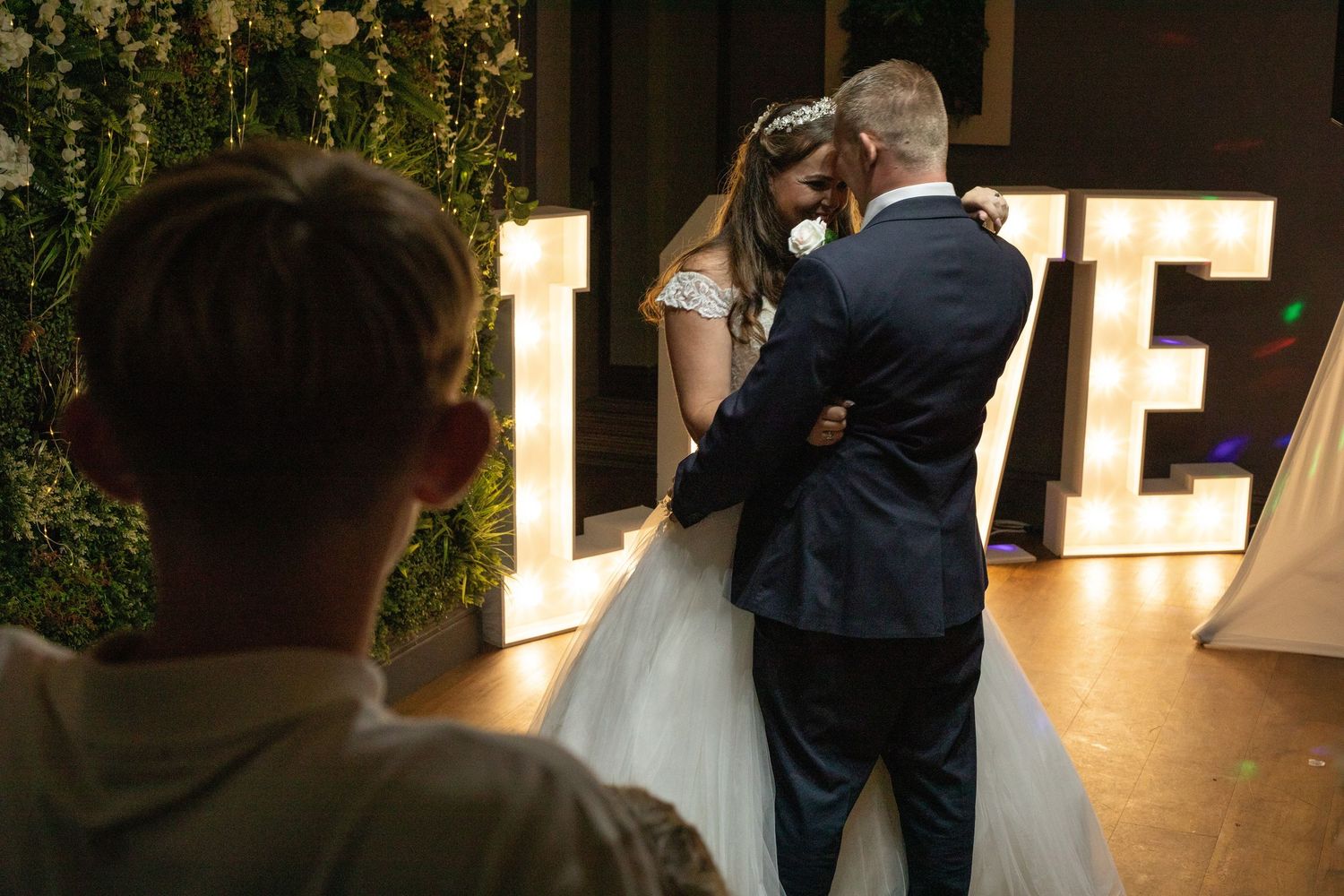 Bride and groom share first dance in front of illuminated LOVE letters at elegant wedding reception.