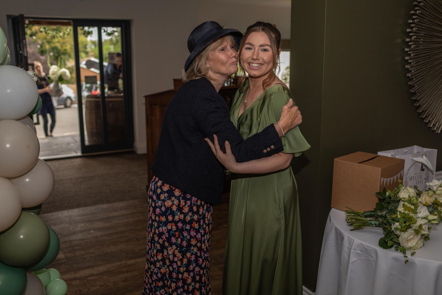 Two women share a warm hug at an elegant event, surrounded by green and white balloon decorations.