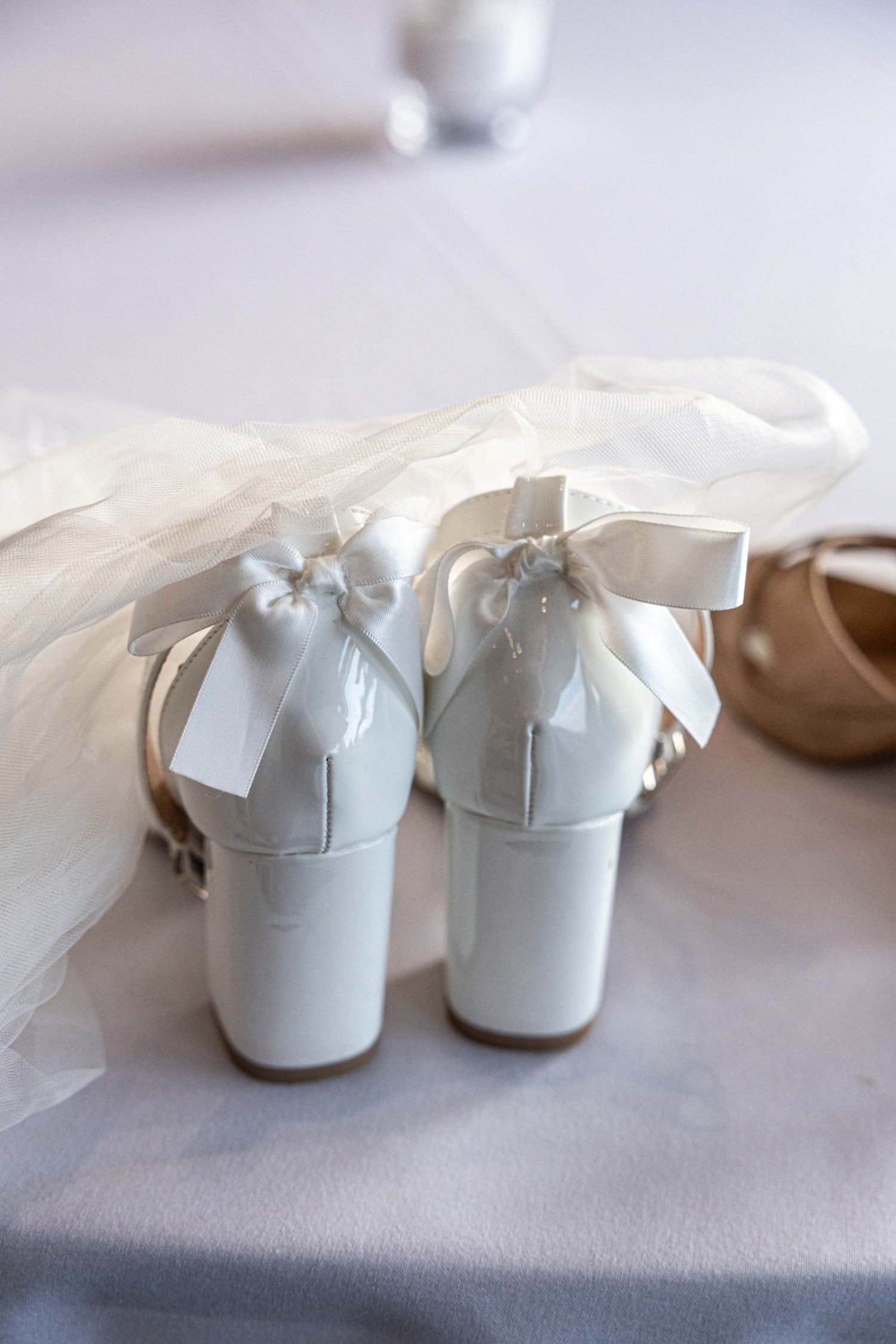 White patent leather bridal heels with satin bows displayed on a white surface with wedding veil.