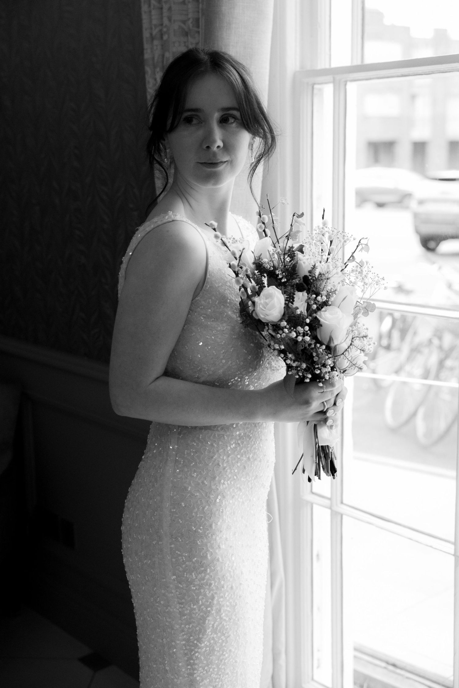 Black and white photo of a bride in a sequined gown holding a floral bouquet while standing by a window.