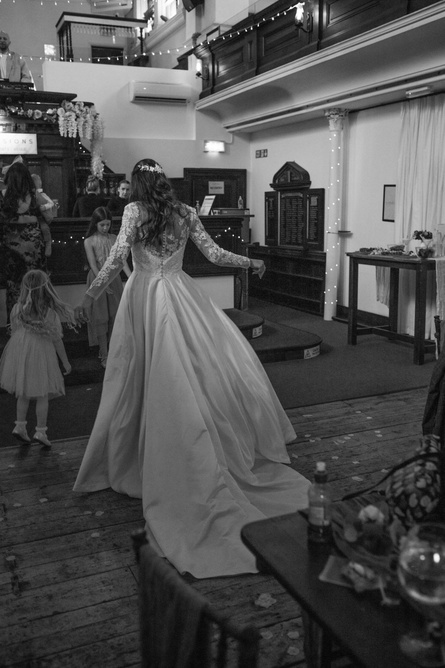 Black and white photo of a bride in a long gown dancing with flower girls in a rustic wedding venue.