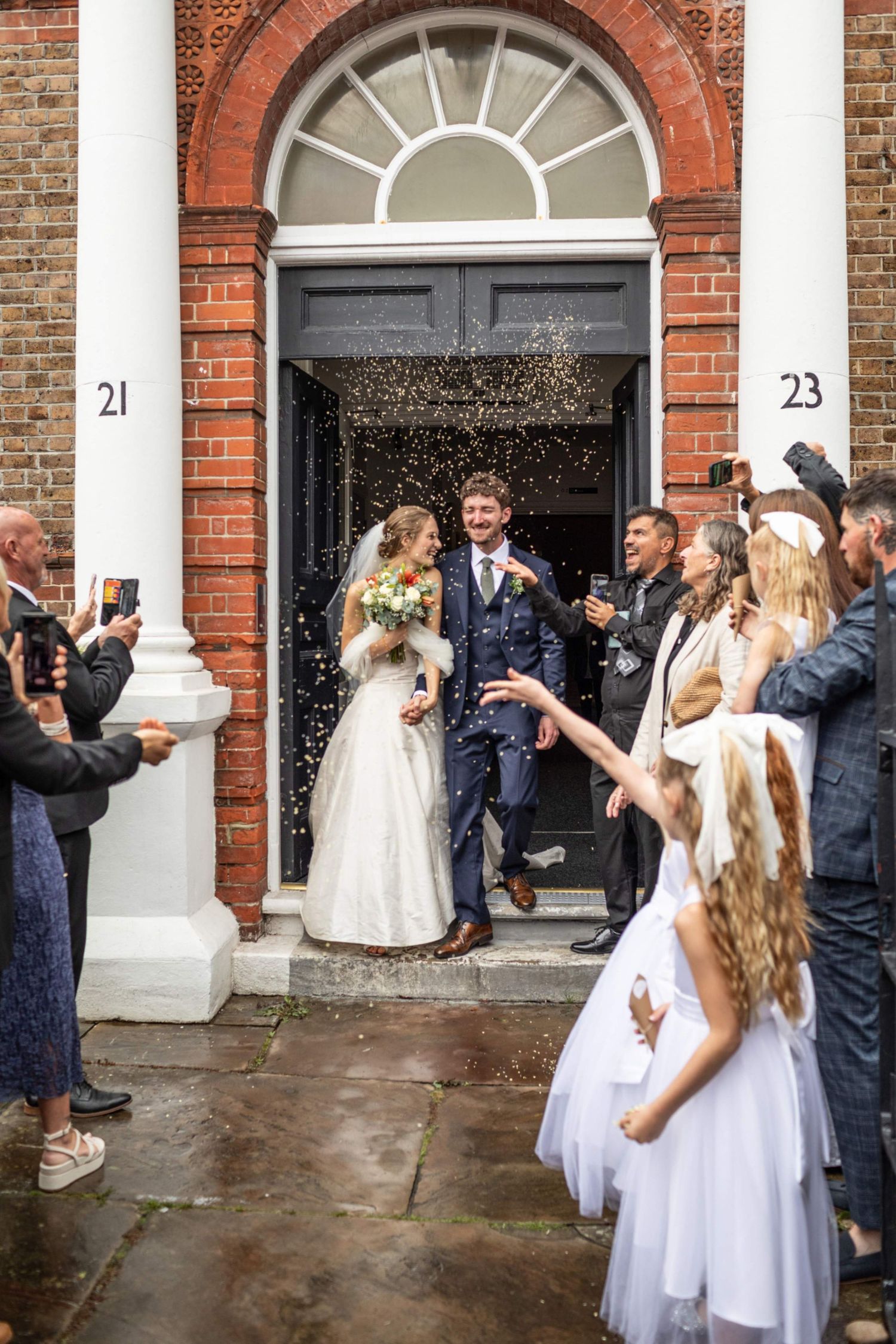 Newlyweds exit brick building as guests celebrate with confetti outside numbered doors 21 and 23.