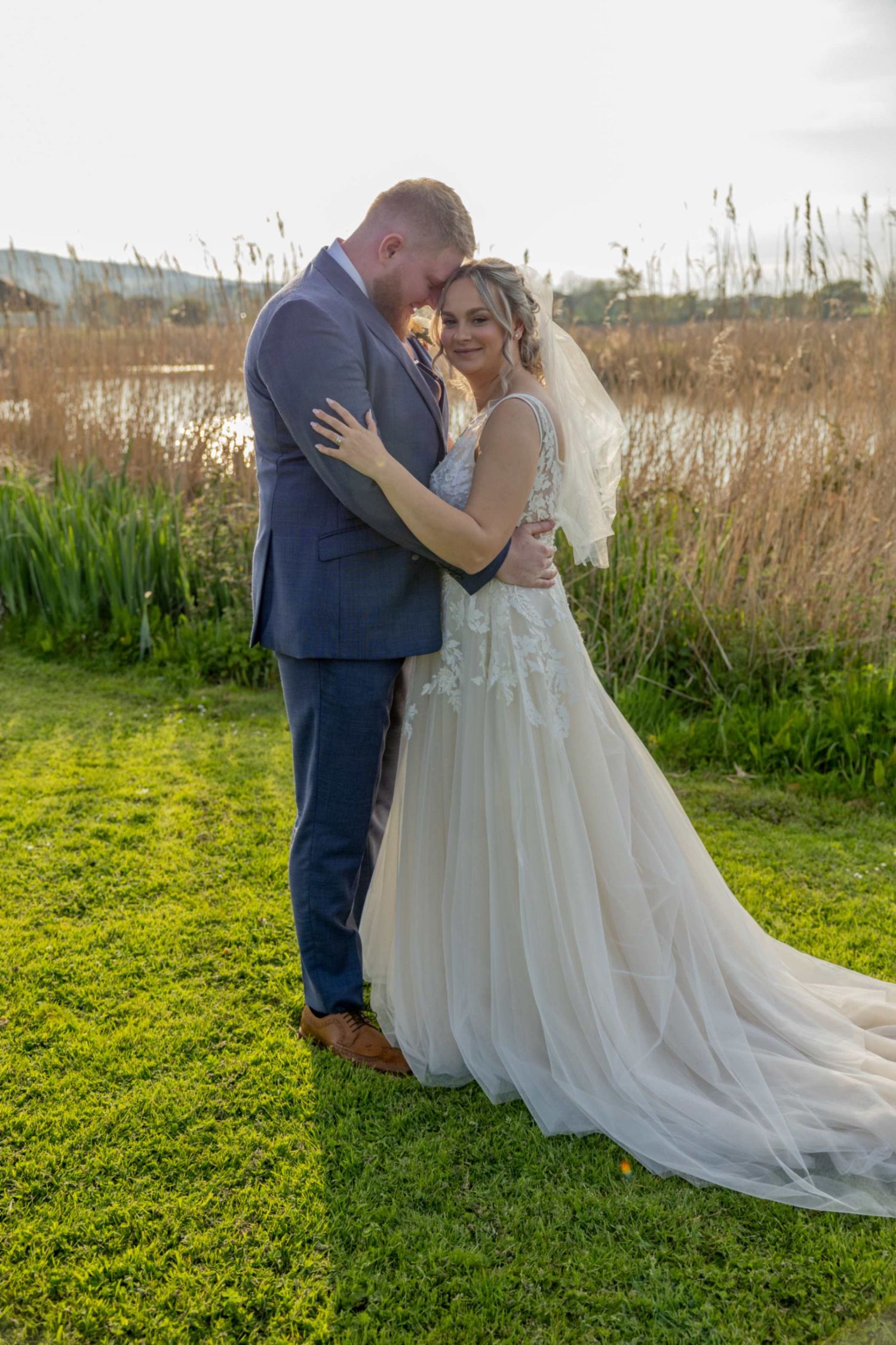 Bride in white gown with veil and groom in blue suit embrace on green grass near a lake at golden hour.