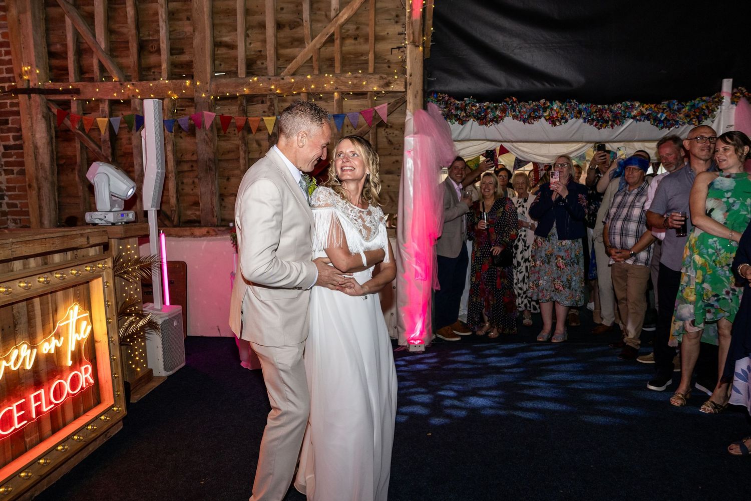Bride and groom share first dance in decorated barn venue with colorful bunting and guests watching at wedding reception.