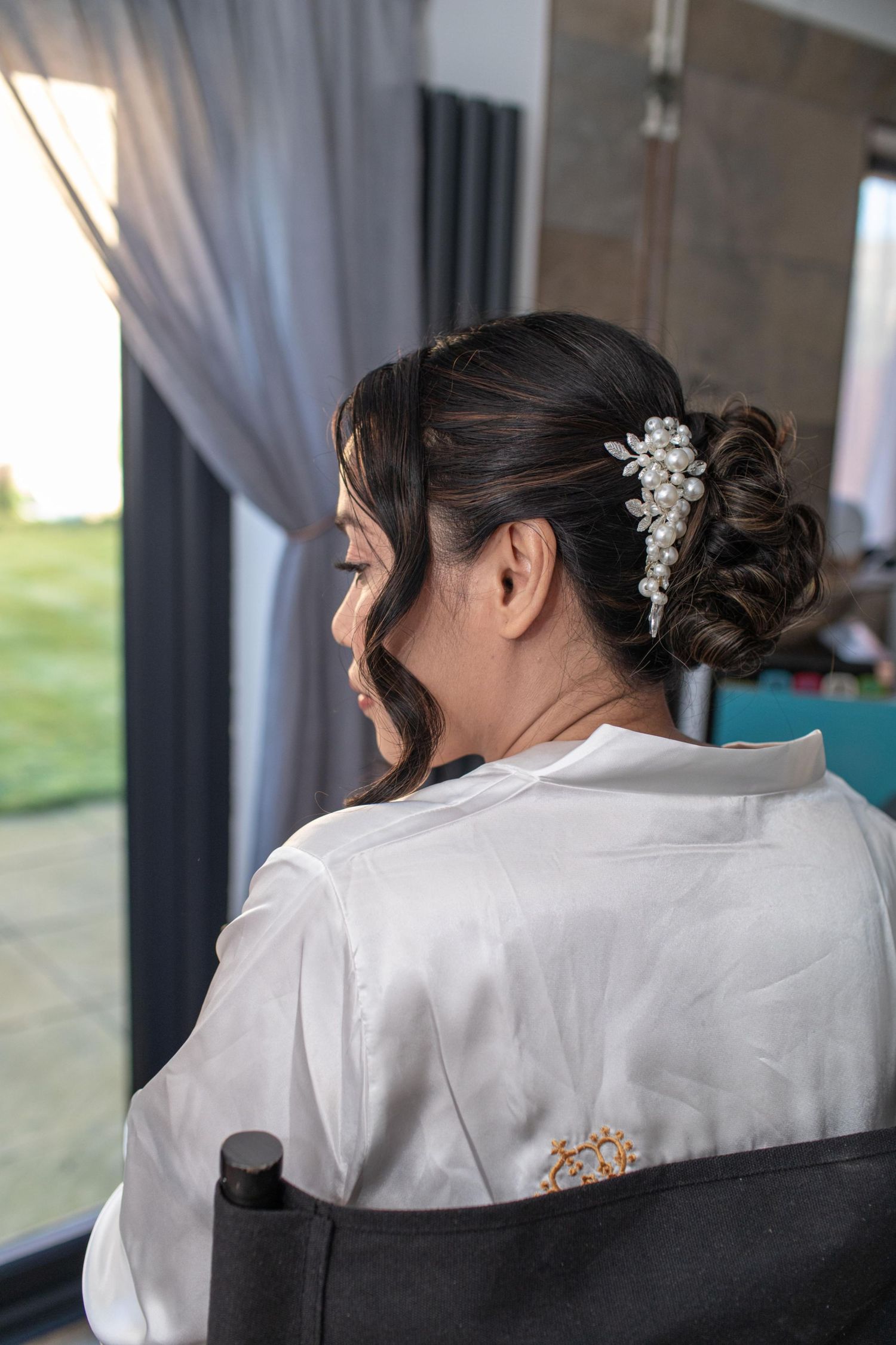 Bride in white robe with elegant updo hairstyle adorned with pearl floral hair accessory, seated before wedding preparation.