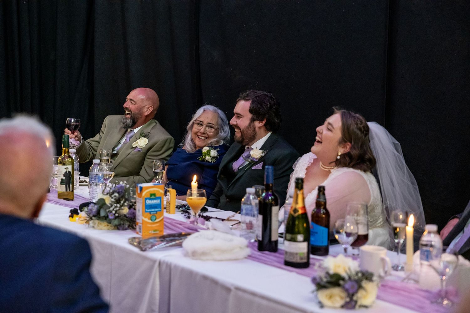 Wedding party laughing at the head table during a reception, with bride in veil and guests enjoying drinks.