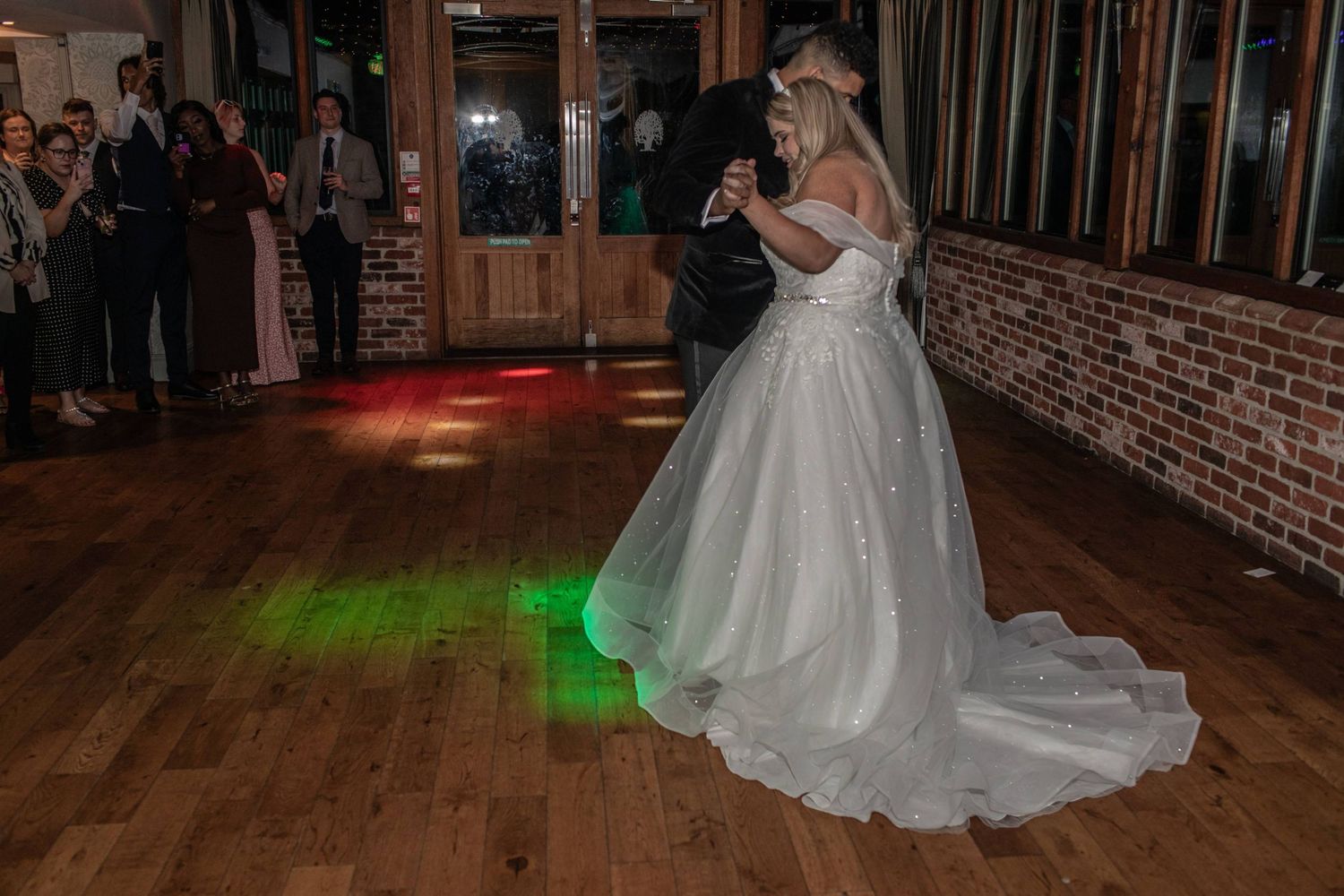 Bride and groom share romantic first dance under green and red lights on wooden dance floor at wedding reception.
