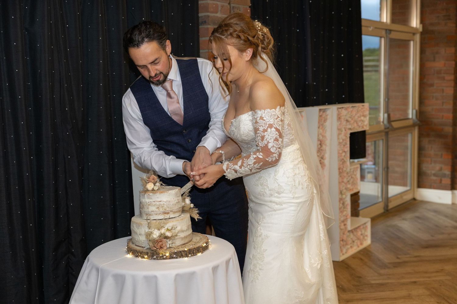 Newlyweds cut their rustic wedding cake together at a wooden barn venue while wearing elegant formal attire.