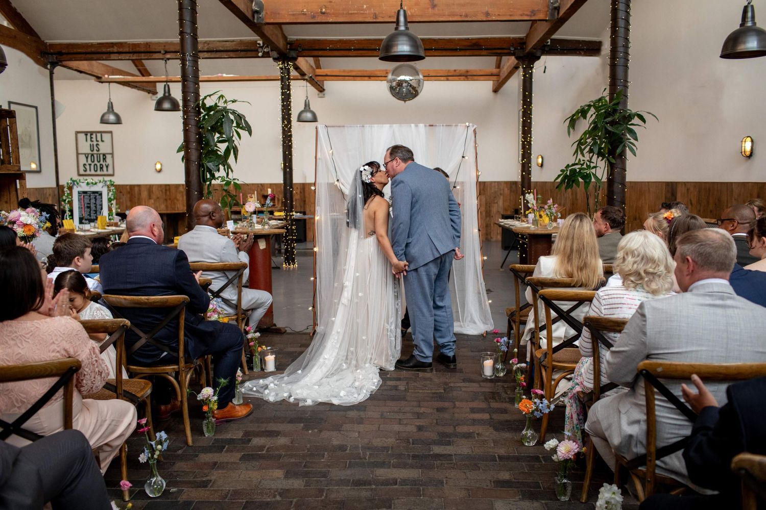 Bride and groom exchange vows at rustic indoor wedding ceremony with white drape backdrop and seated guests.