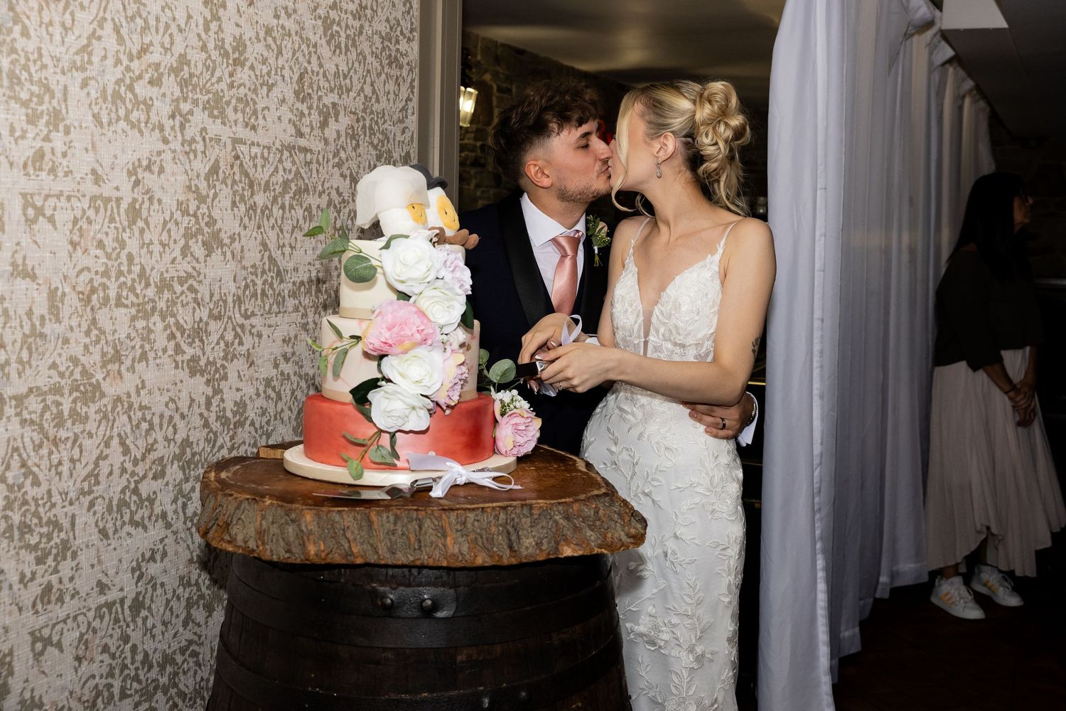 Bride and groom kissing while cutting a floral white wedding cake displayed on a rustic wooden barrel.