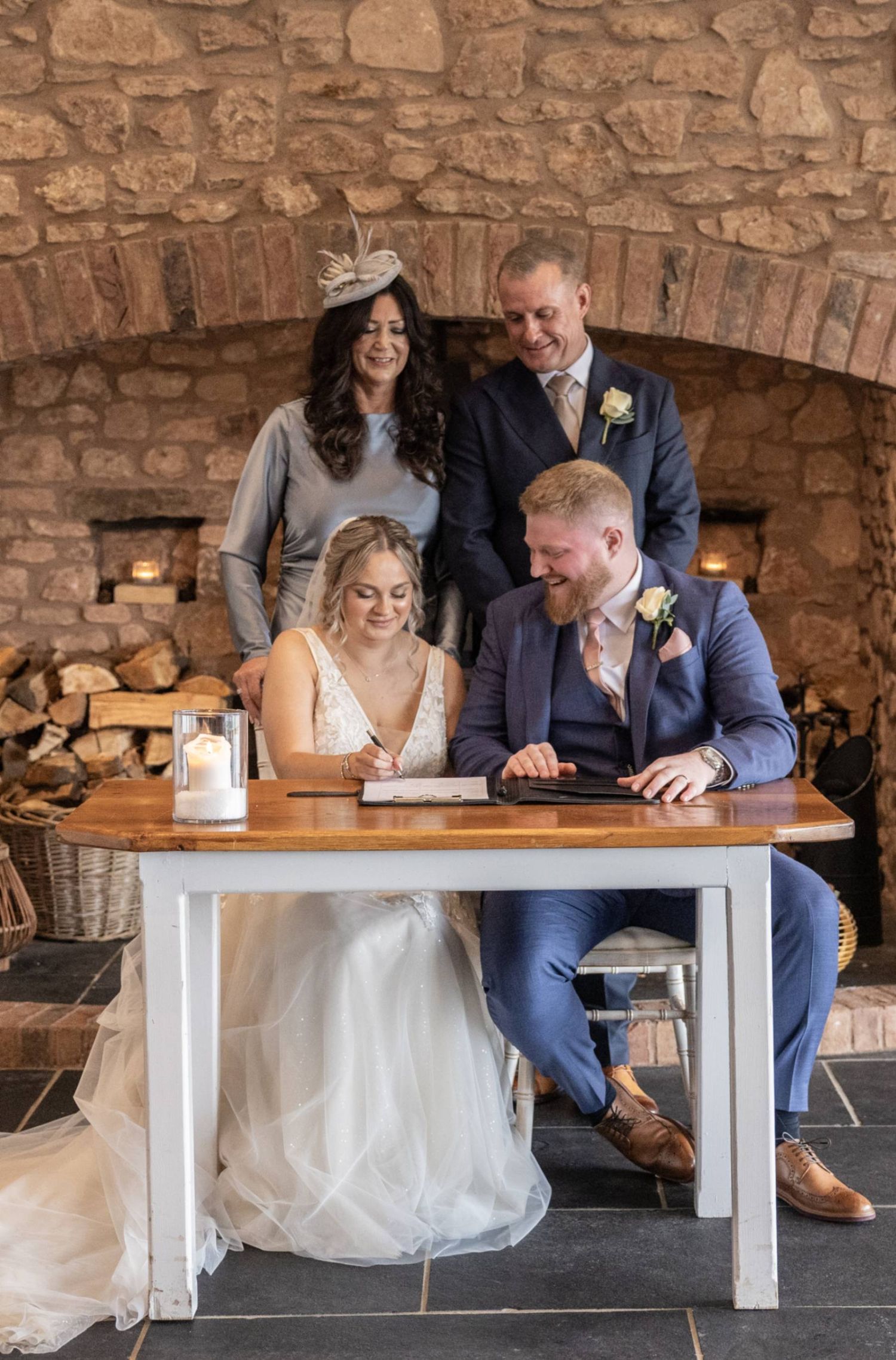 Bride and groom sign wedding register at rustic stone venue, witnessed by two smiling guests standing behind them.