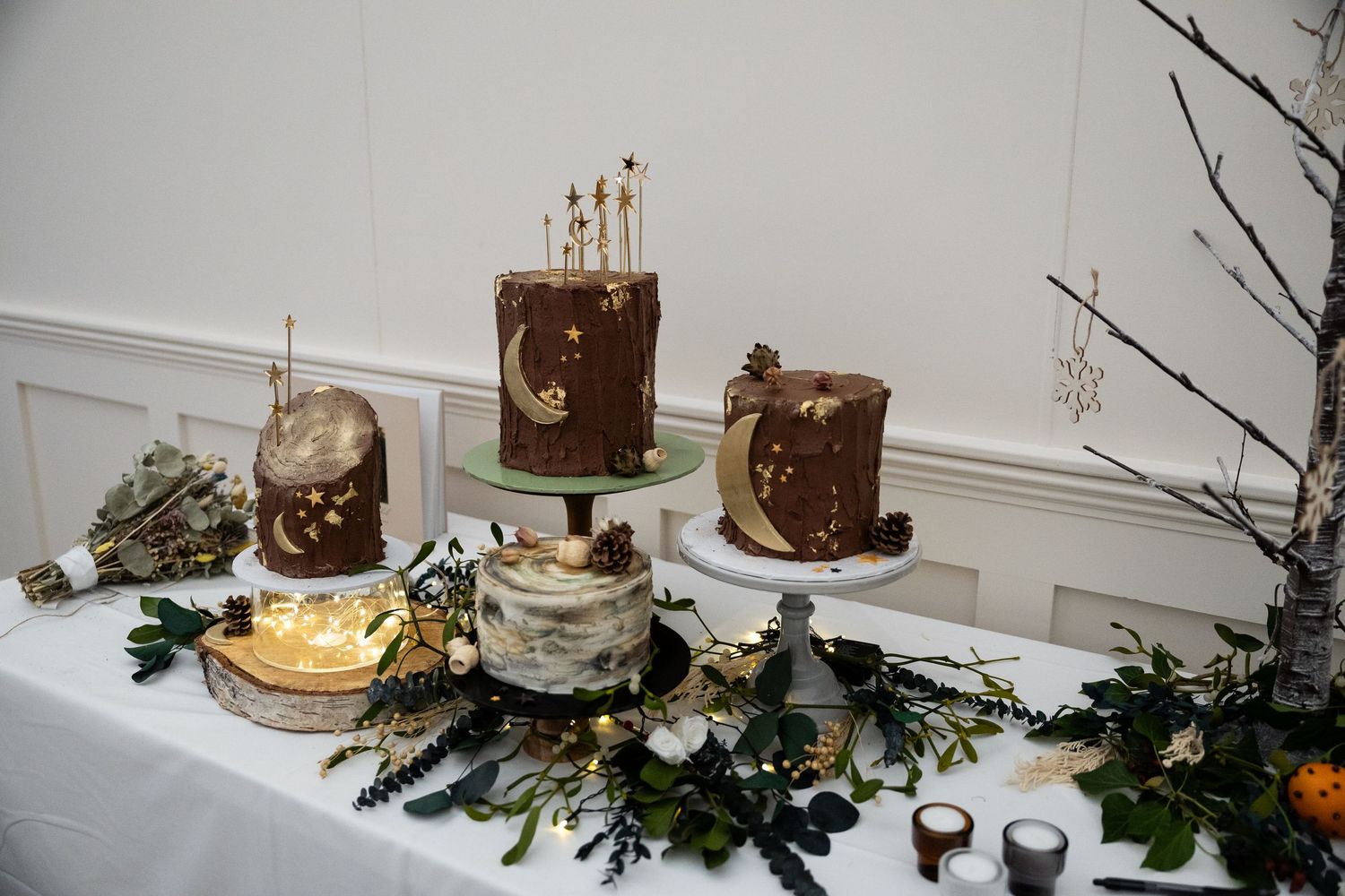 A collection of rustic chocolate and vanilla cakes decorated with greenery and candles on a white table display.