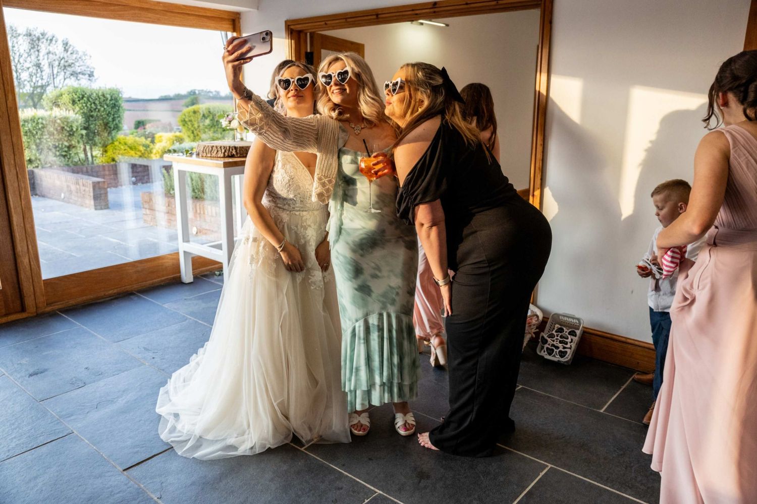 Bride takes a selfie with guests wearing sunglasses during wedding preparations indoors.