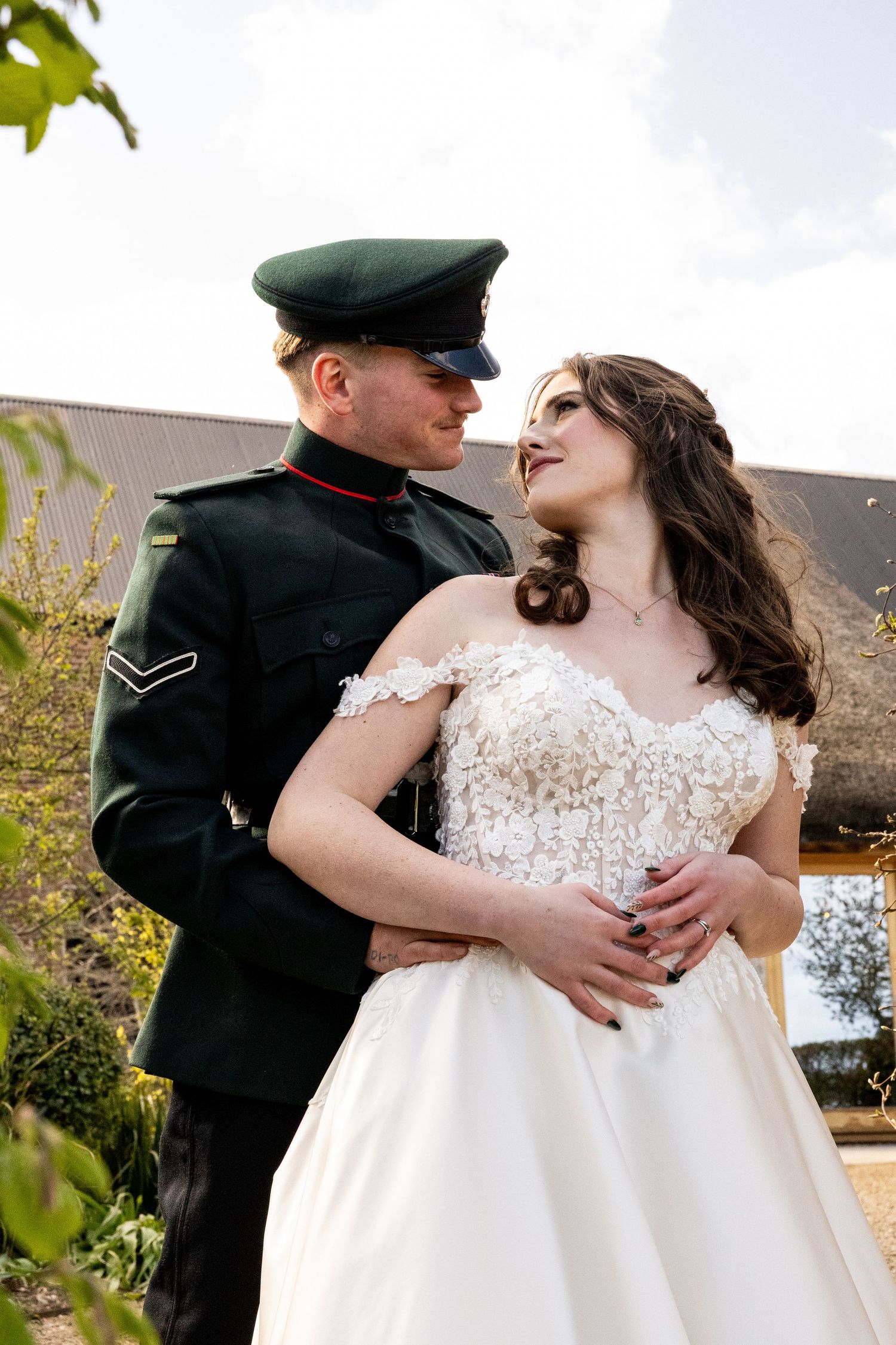 Military groom in green uniform embraces bride in lace wedding dress outdoors at rustic venue.