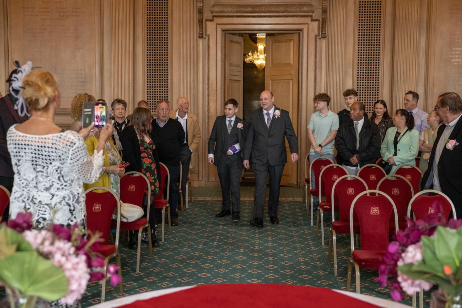 Two grooms walk down the aisle at an elegant wedding ceremony with red chairs and green carpet.