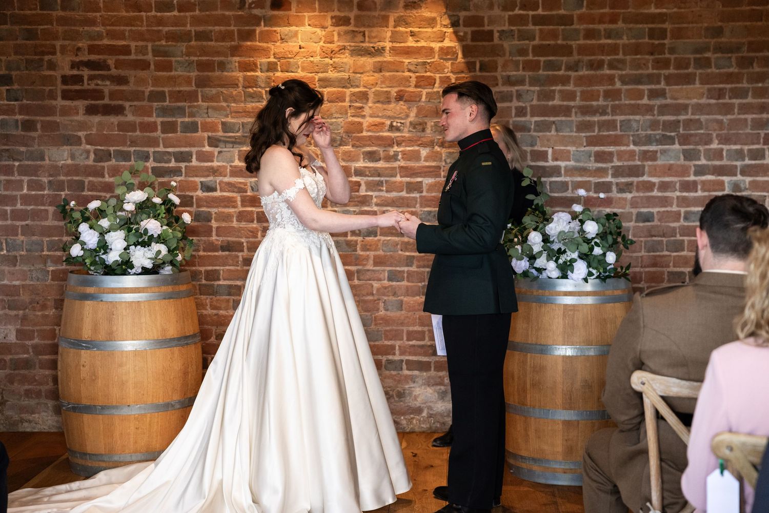 Bride and groom exchange vows at rustic brick wall wedding ceremony with white floral arrangements on wooden barrels.