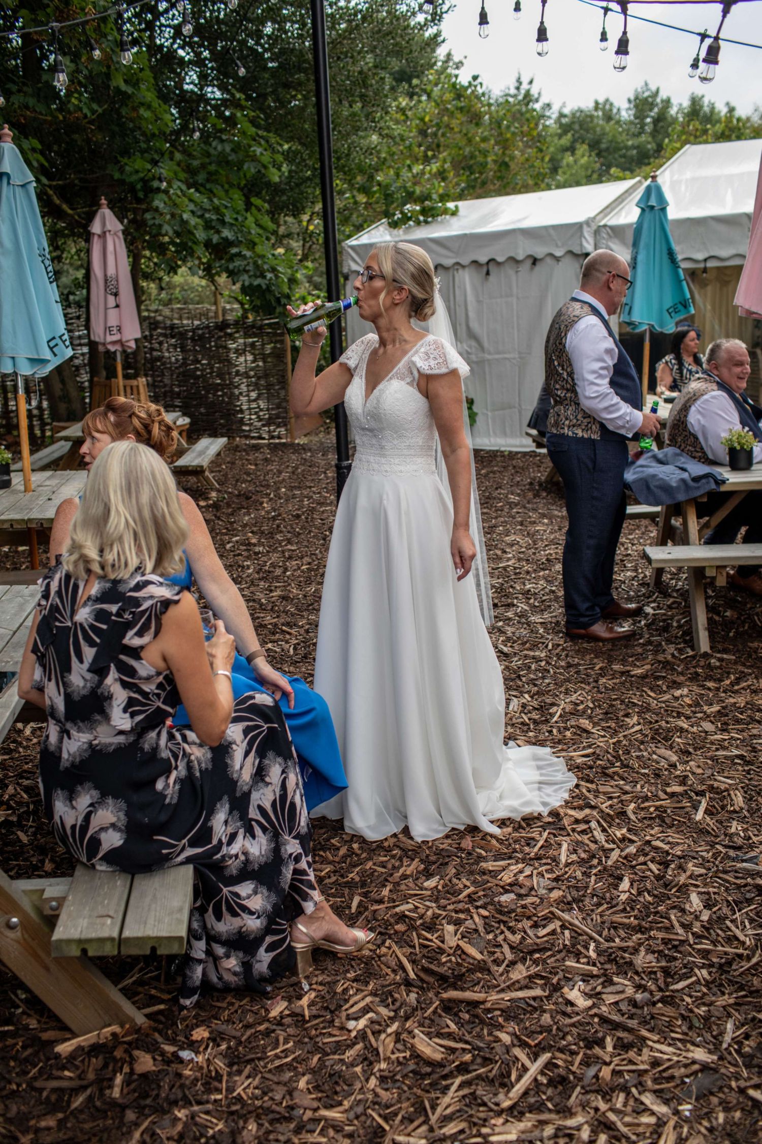 Bride in white gown drinks at outdoor wedding reception with guests on wood chip ground near marquee tent.