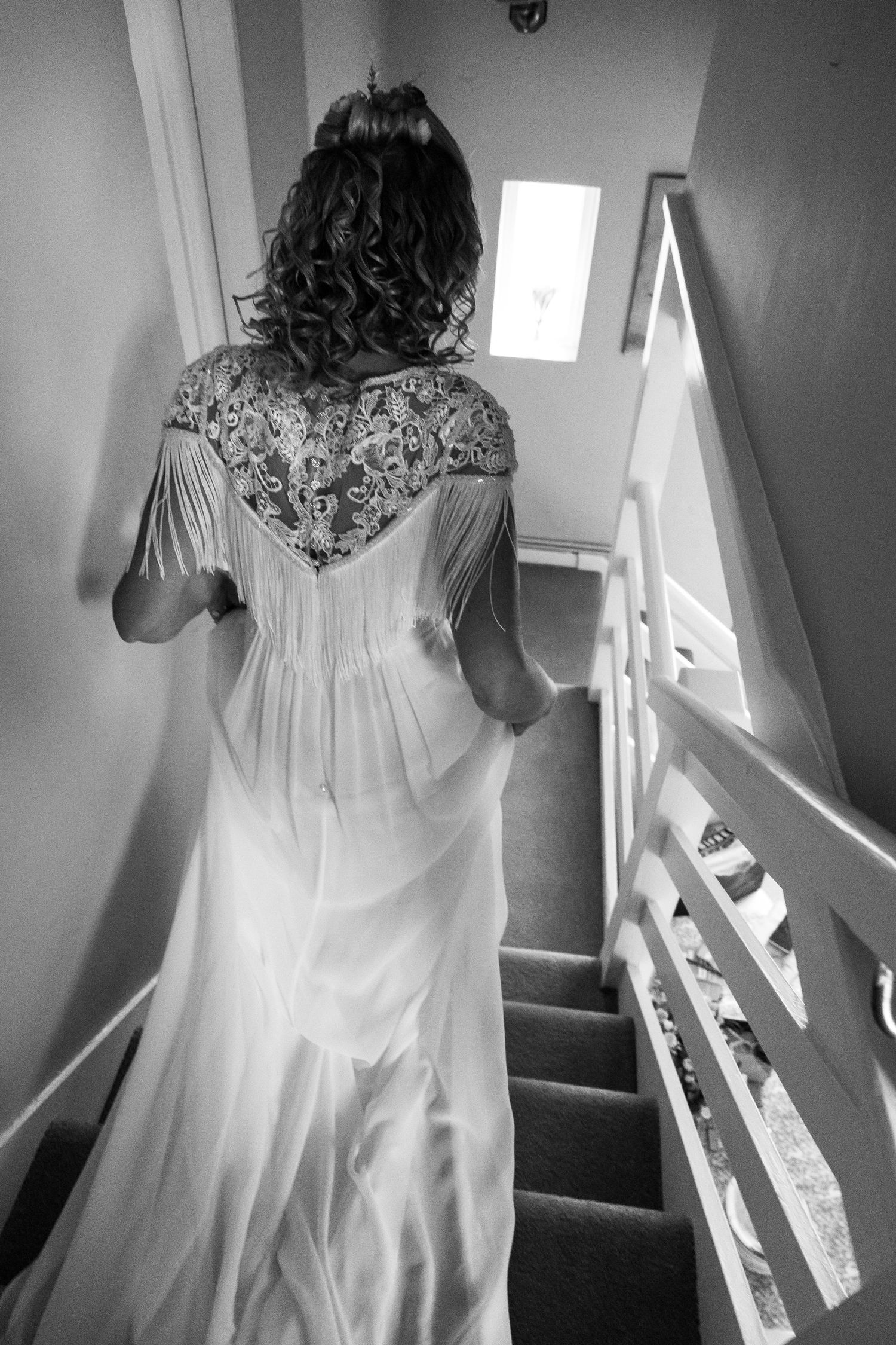 Black and white photo of bride in lace wedding dress ascending staircase, showing intricate back detail.