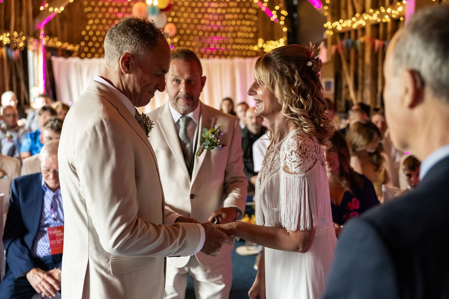 Couple exchanging rings during an outdoor barn wedding ceremony with guests seated in background.