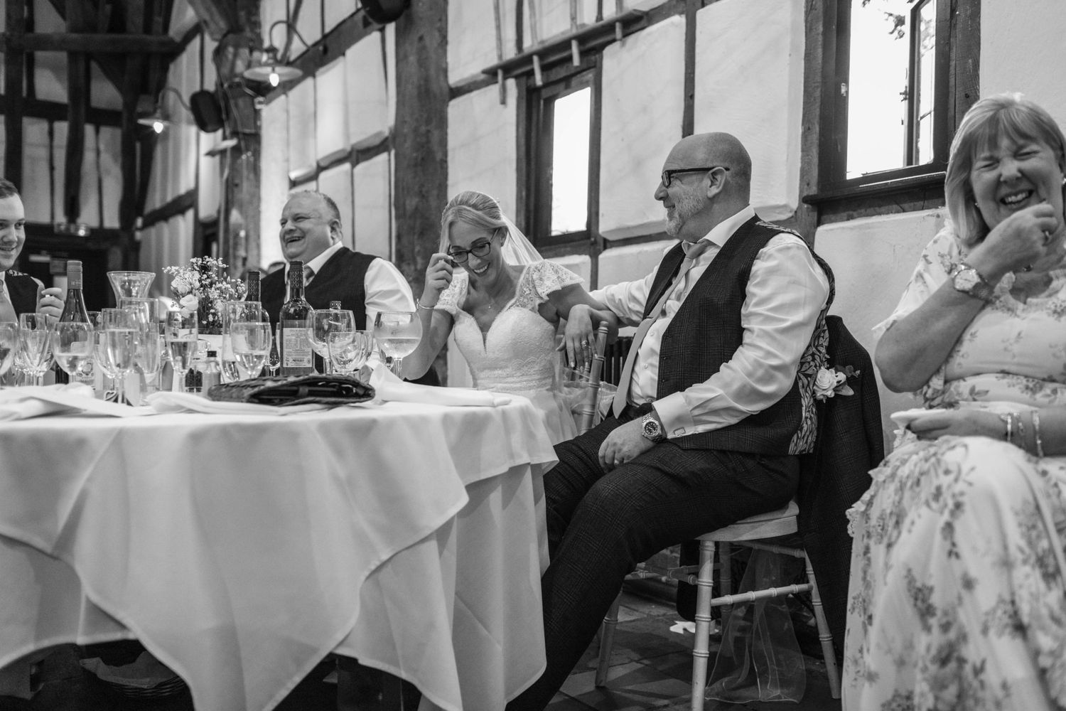 Black and white wedding reception photo showing guests laughing at the head table in a rustic venue.