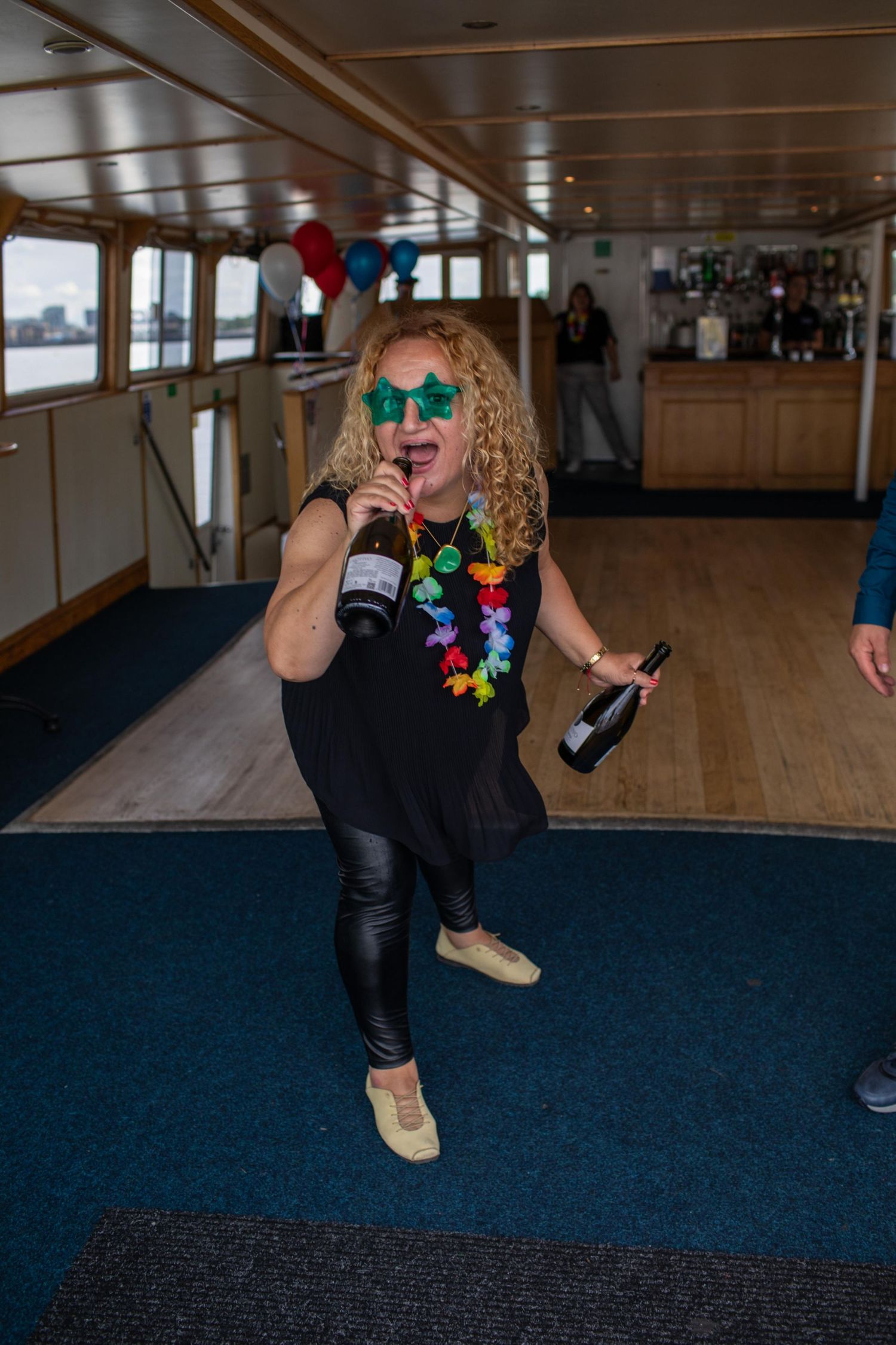 Woman dancing on a boat party, wearing green star sunglasses, floral lei, holding champagne bottles.