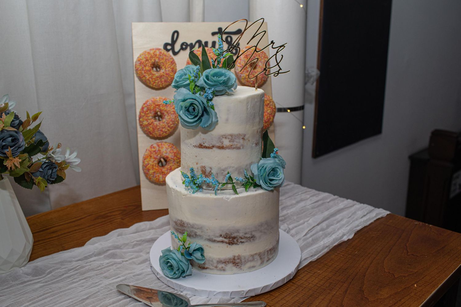 Two-tier semi-naked wedding cake with teal roses beside a donut wall display on a wooden table.