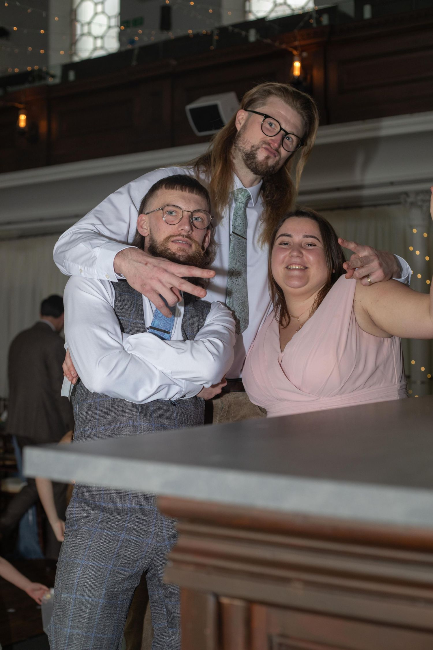 Three friends pose together at a wedding reception, smiling at the bar in formal attire.